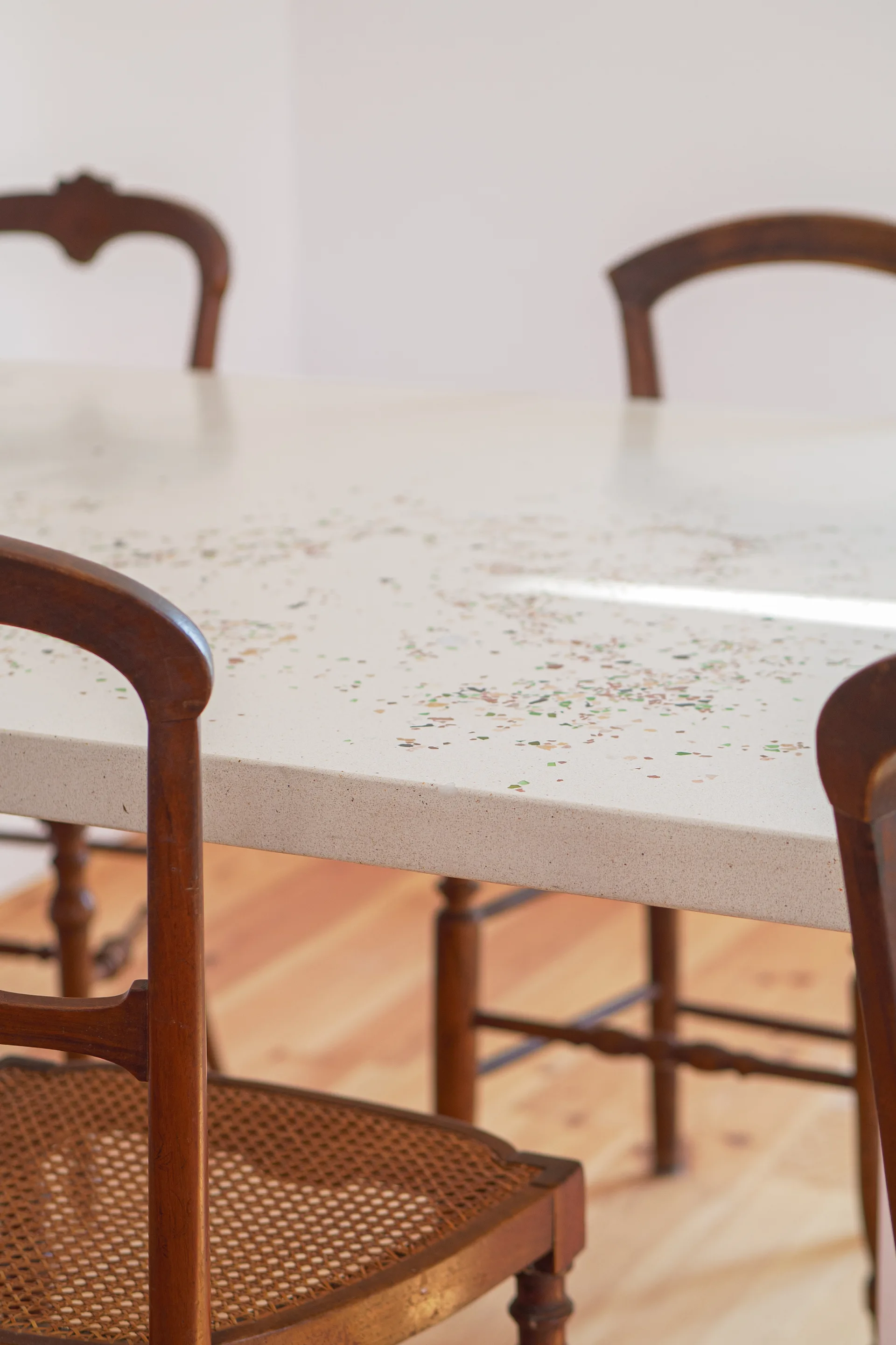 A speckled off-white terrazzo dining table is paired with traditional wooden chairs featuring woven cane seats on a light wood floor.
