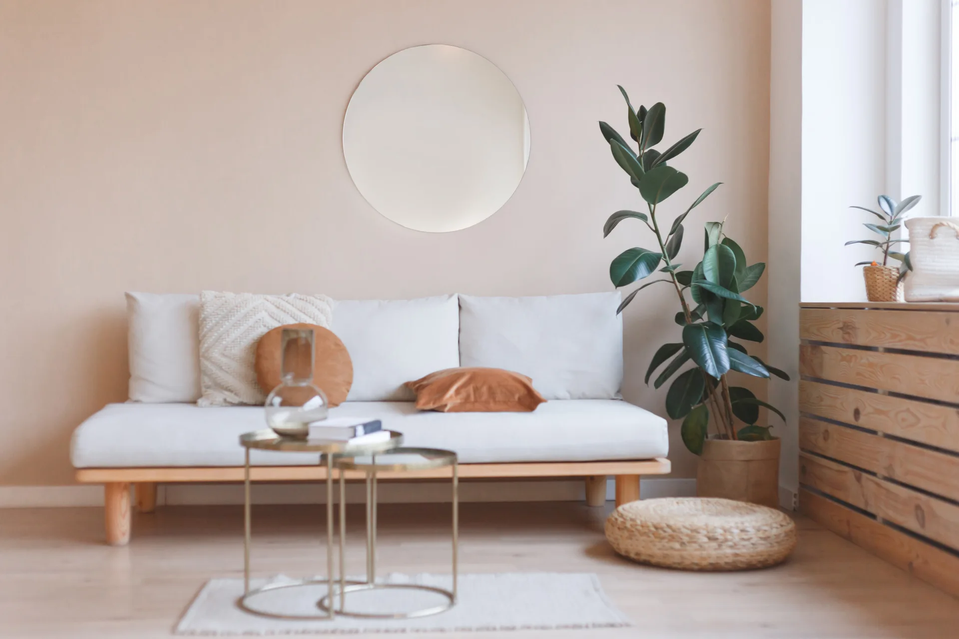 A minimalist beige living room features a white daybed sofa with brown cushions under a round wall mirror, flanked by a potted rubber plant.