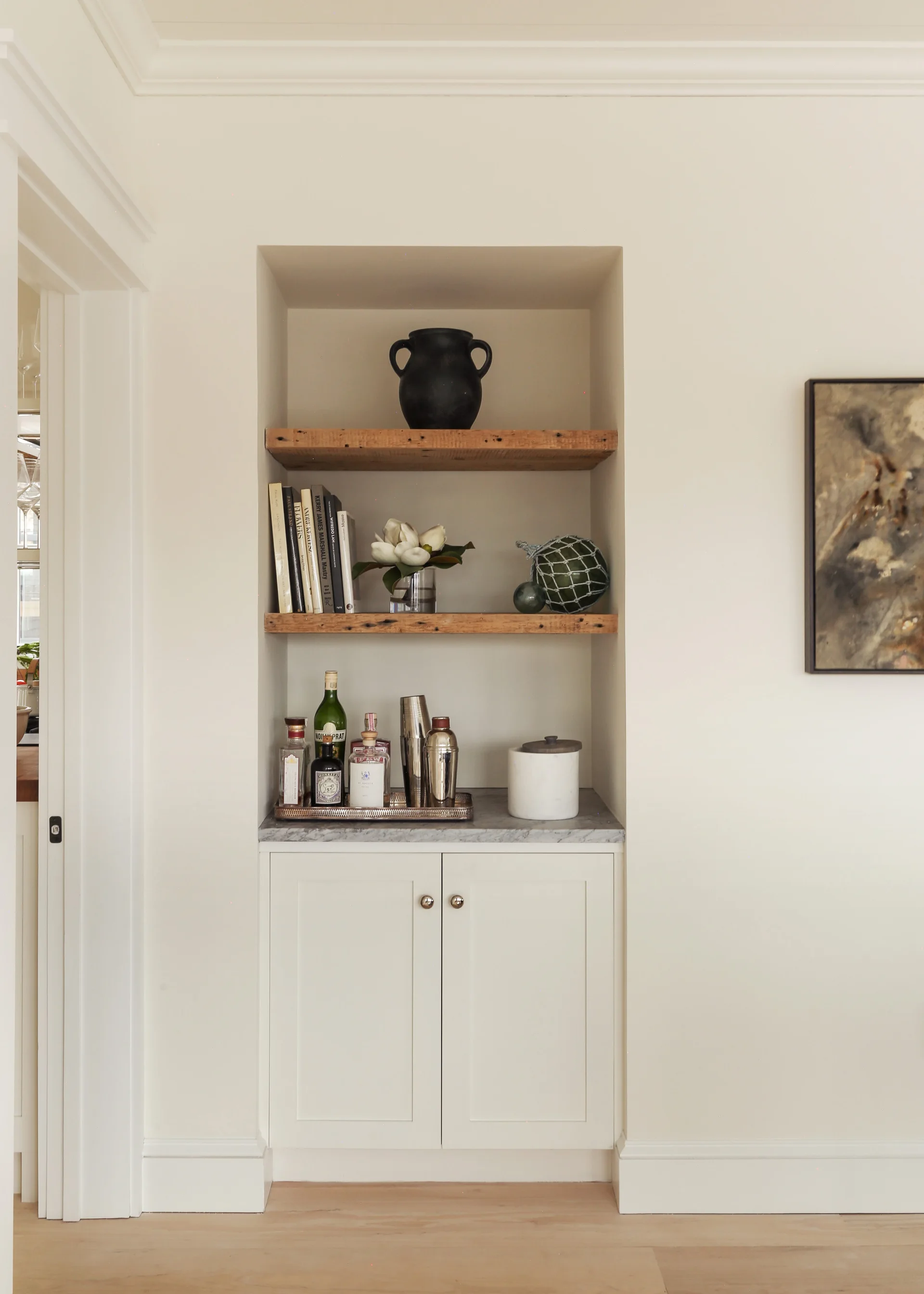 A built-in white bar cabinet recessed in a wall niche features a marble countertop and two rustic wood floating shelves styled with books and art objects.