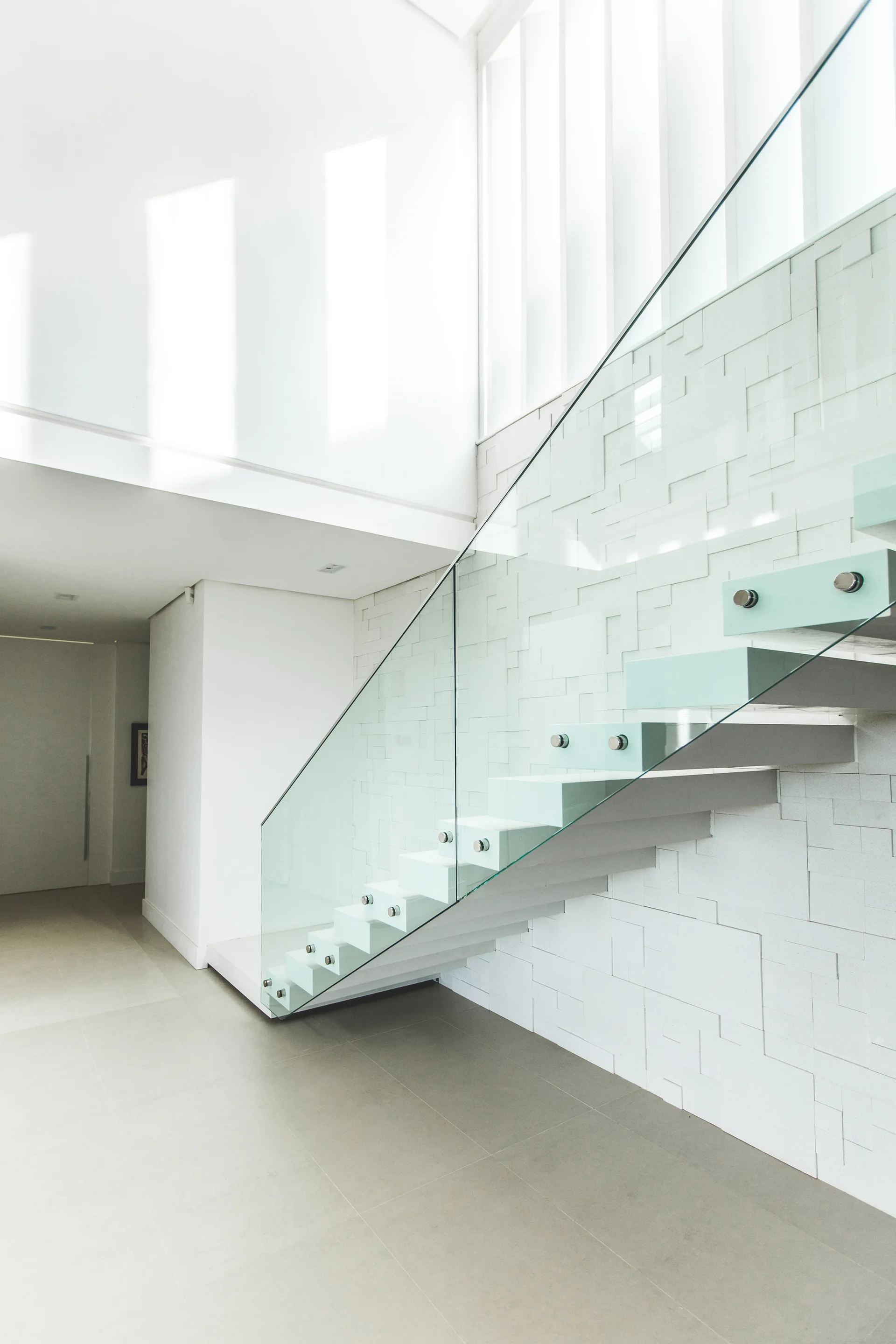 A minimalist white floating staircase with a clear glass railing stands against a geometric textured tile wall in a bright modern home.