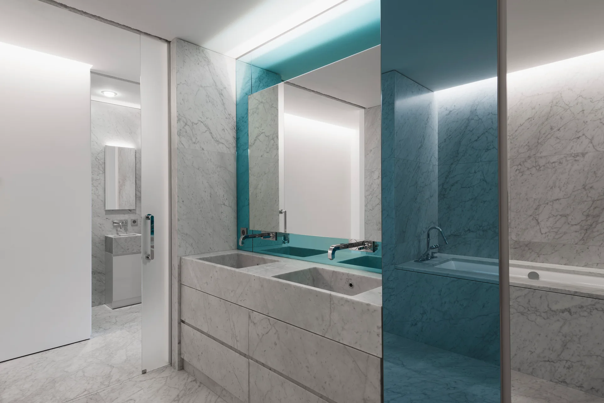 A minimalist bathroom featuring grey-veined white marble walls and floors, a double vanity with integrated sinks, and a large blue-tinted mirror.