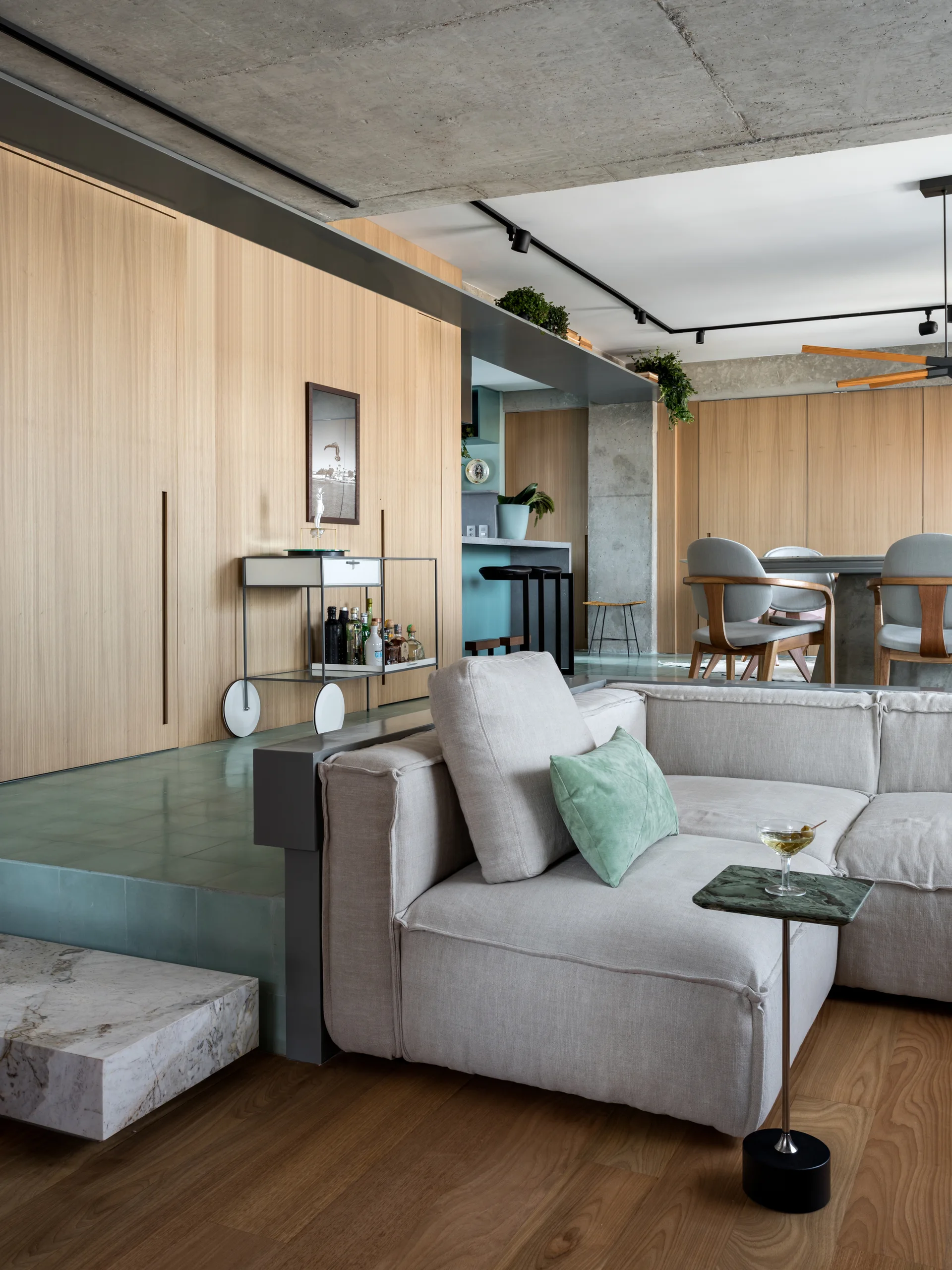 A low grey sectional sofa on a hardwood floor sits below an elevated platform tiled in green, backed by tall wood wall paneling.