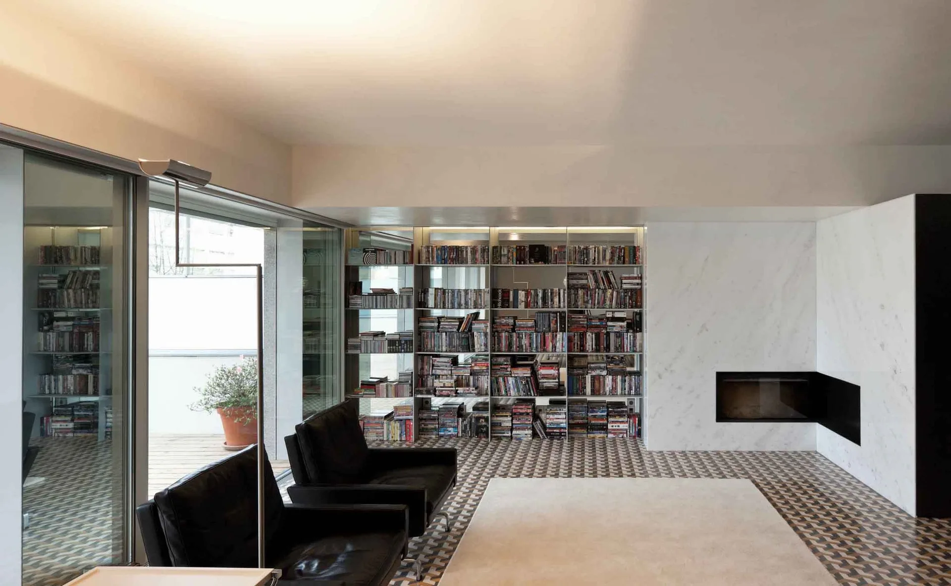 A modern living room features two black leather lounge chairs on a geometric tiled floor next to a white marble fireplace and a recessed metal bookshelf.