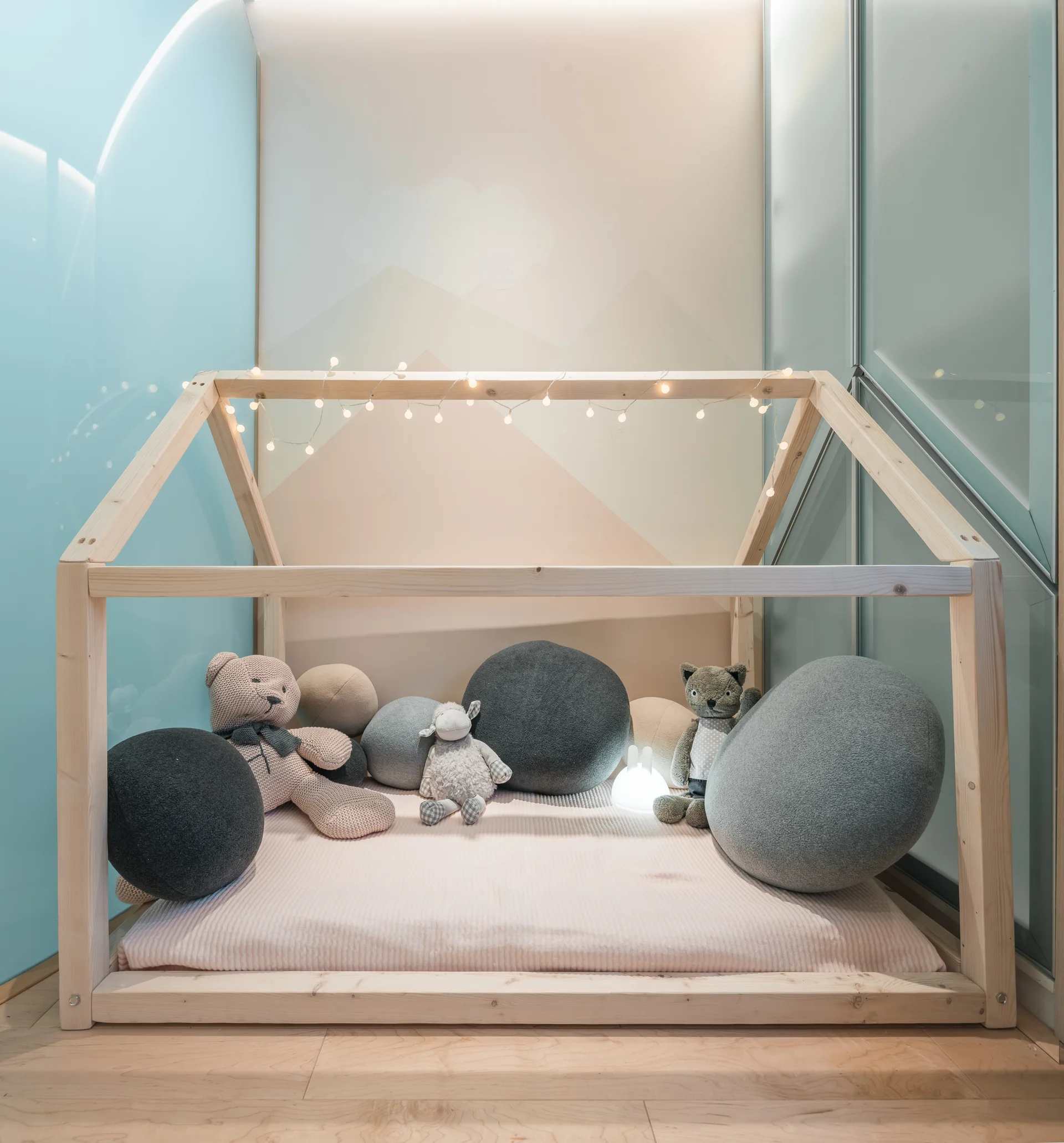 A children's wooden house-shaped bed frame sits on a light wood floor, decorated with round plush cushions, stuffed toys, and warm string lights.