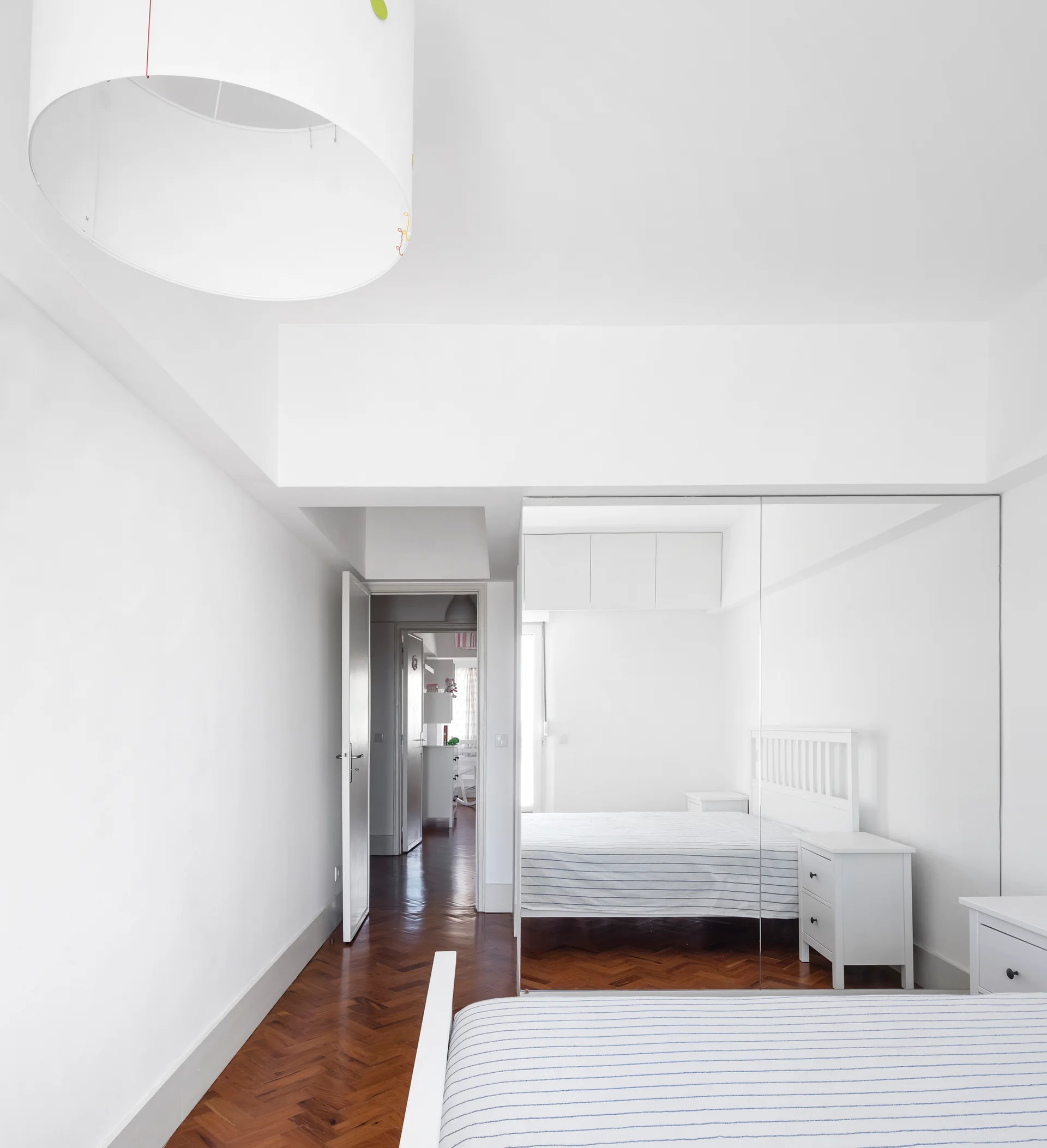 A minimalist white bedroom featuring dark herringbone parquet floors, a bed with striped bedding, and floor-to-ceiling mirrored closet doors.
