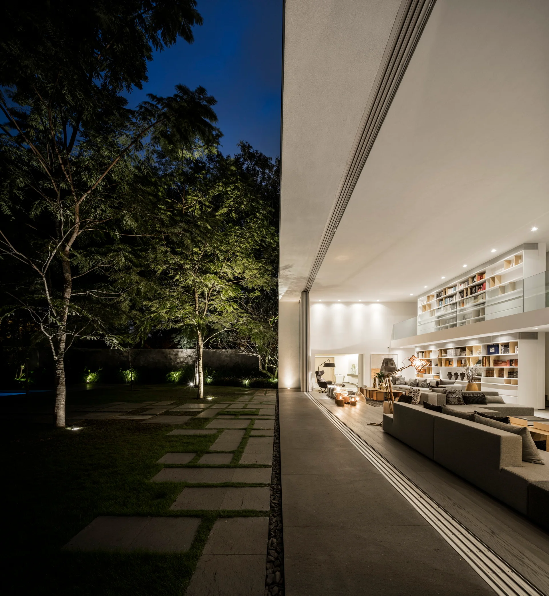 A modern living room with a large gray sectional sofa and a two-story bookcase opens directly onto an illuminated outdoor patio and garden.