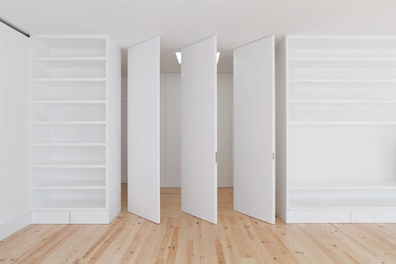 White floor-to-ceiling built-in shelving units flank three white pivoting door panels over a light natural wood plank floor.