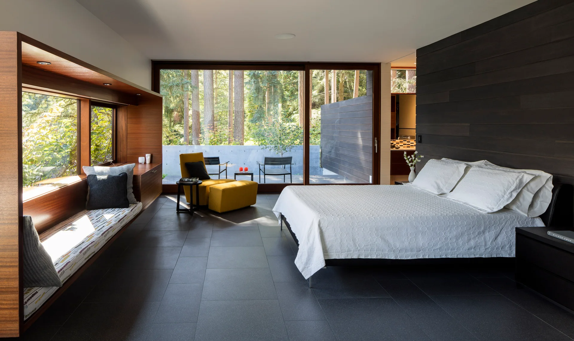 A modern bedroom features a large bed with white bedding, a built-in wood window seat, a mustard yellow lounge chair, and sliding glass patio doors.