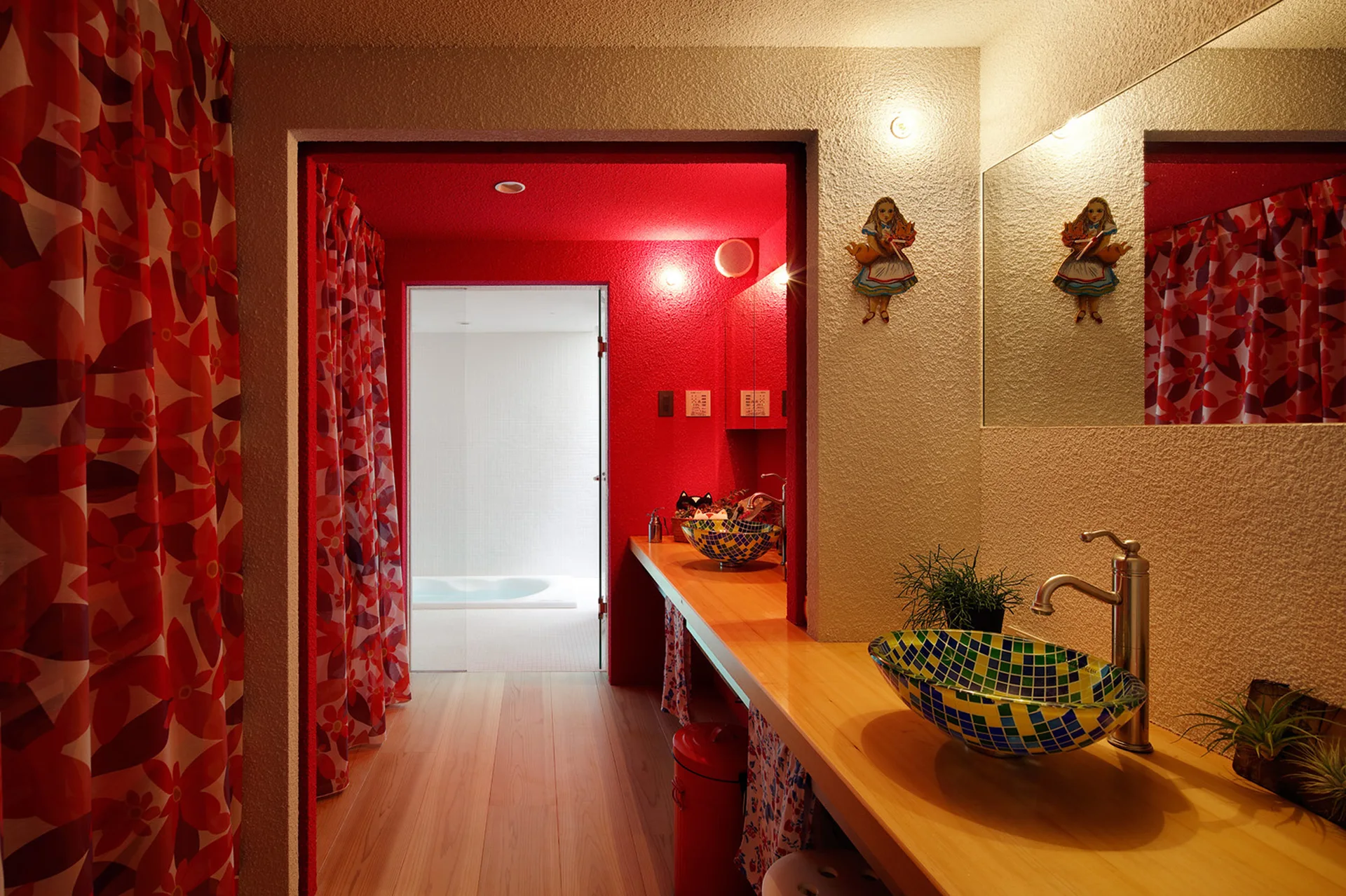 A wooden vanity counter holds a mosaic glass vessel sink beneath a mirror, set against textured walls and a vibrant red doorway.