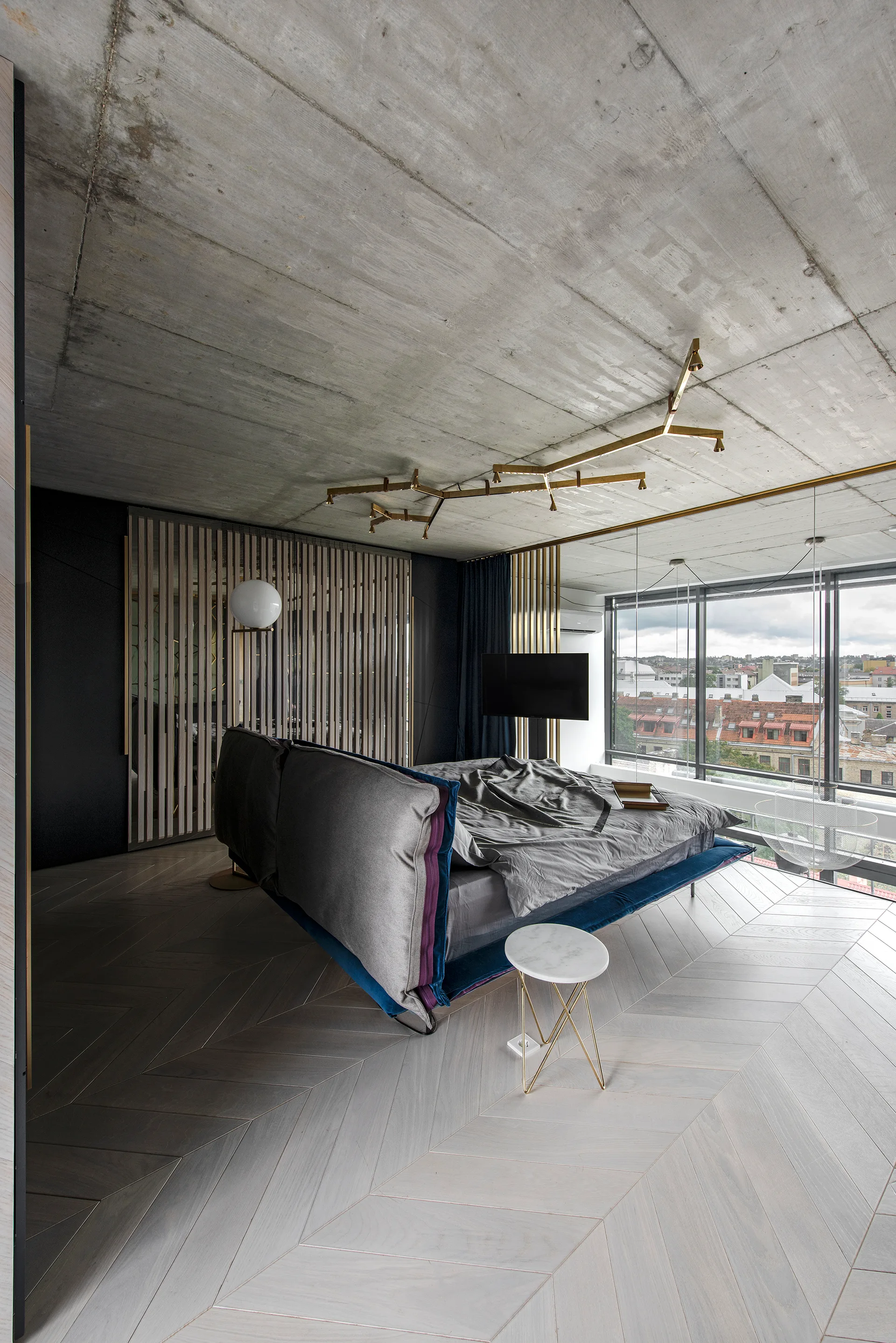 A modern bedroom features a low-profile bed, light grey herringbone wood floors, a raw exposed concrete ceiling, and a thin branched gold light fixture.