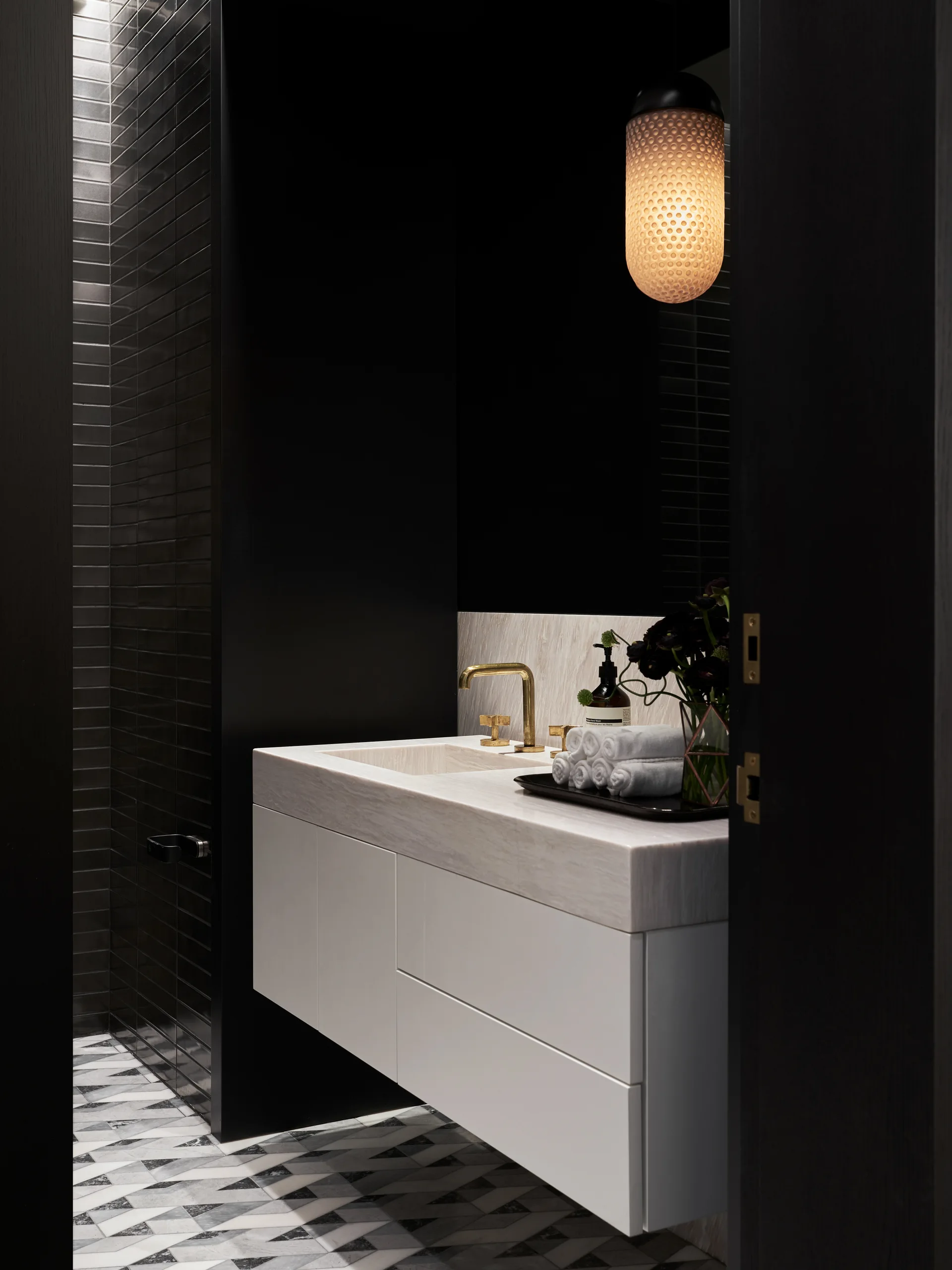 A modern powder room featuring a white floating vanity with a marble countertop, gold faucet, and a black wall with geometric floor tiles.