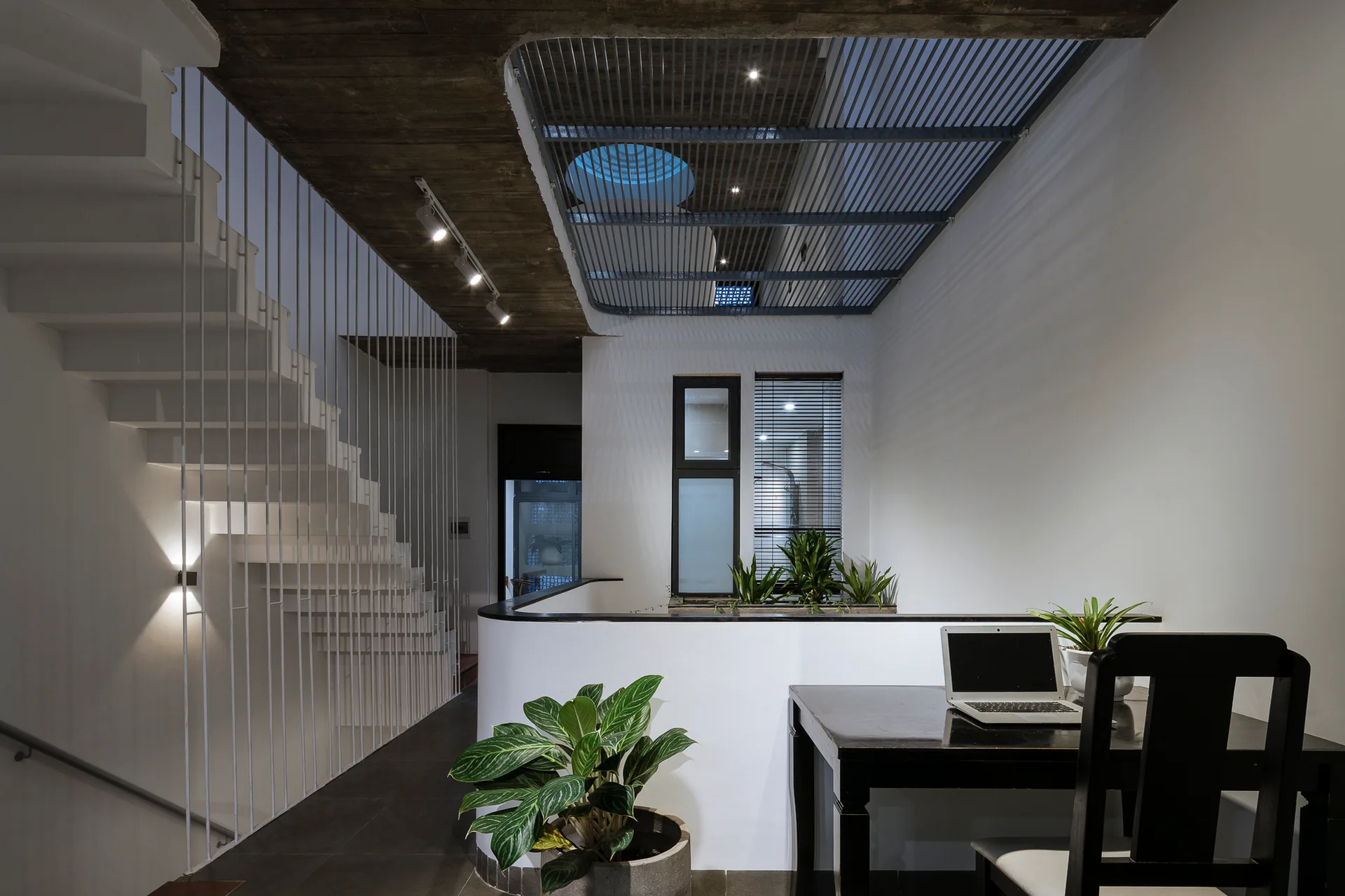 A minimalist mezzanine landing featuring a white staircase with a vertical metal spindle screen, a black wooden desk, and a metal ceiling grid.