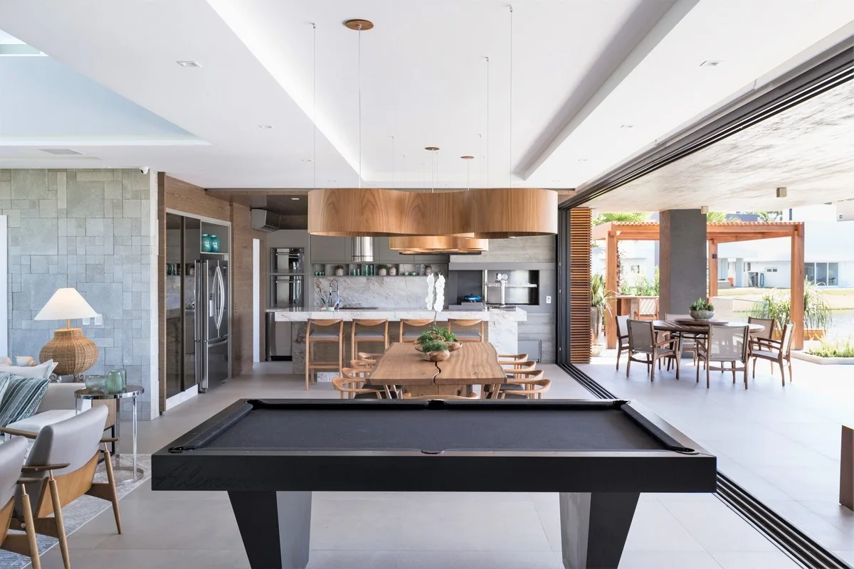 An open-concept living area features a black pool table, a live-edge wooden dining table beneath curved wood pendant lights, and sliding glass doors.