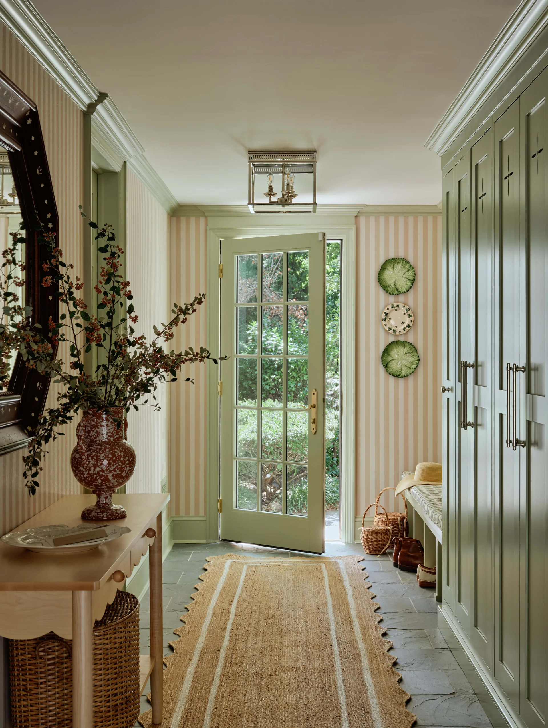 A traditional entryway features sage green built-in cabinets, striped wallpaper, a scalloped jute runner over slate flooring, and an open glass door.
