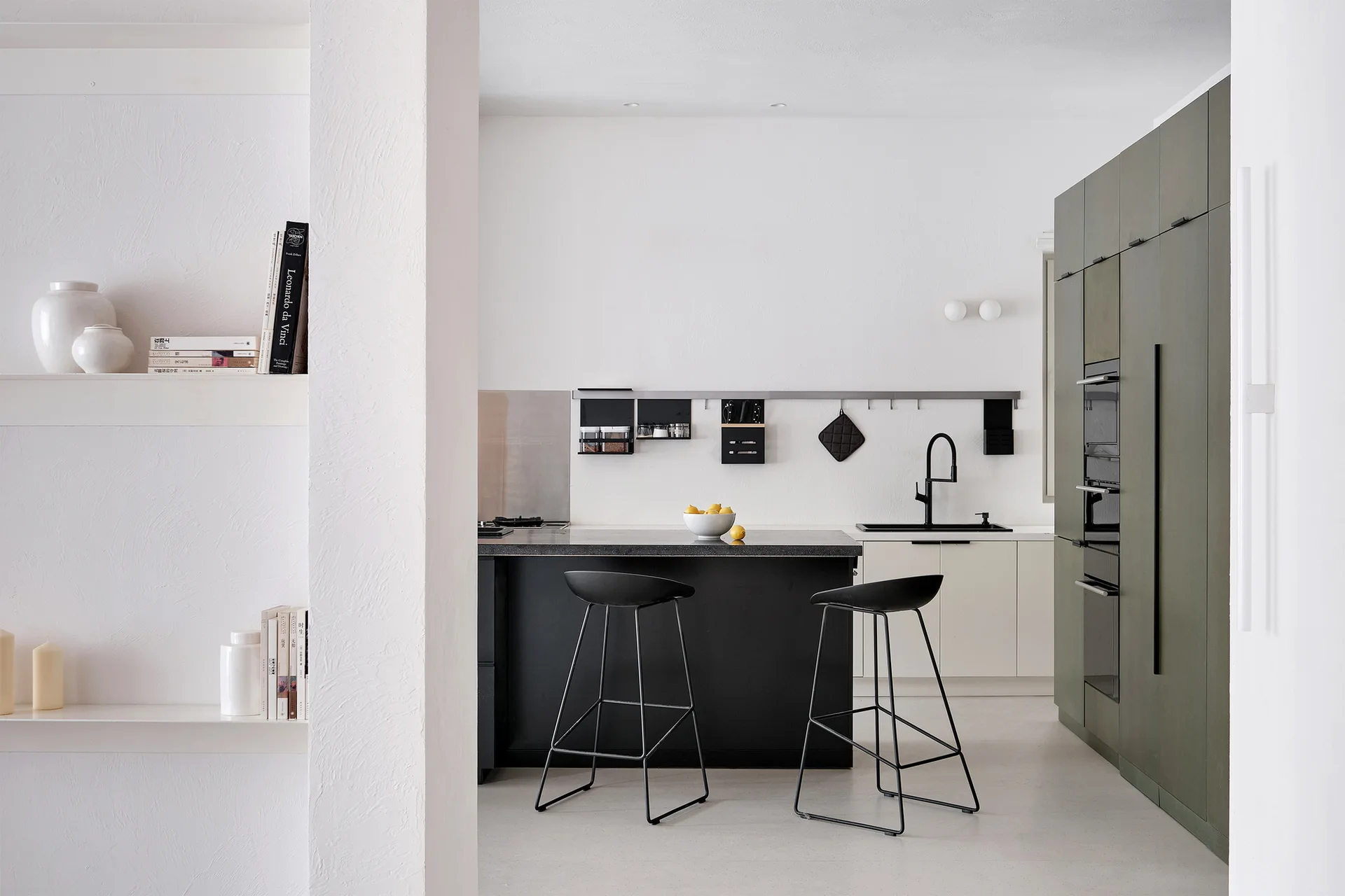 A minimalist kitchen features matte olive green tall cabinets, a black island with two bar stools, and textured white walls.