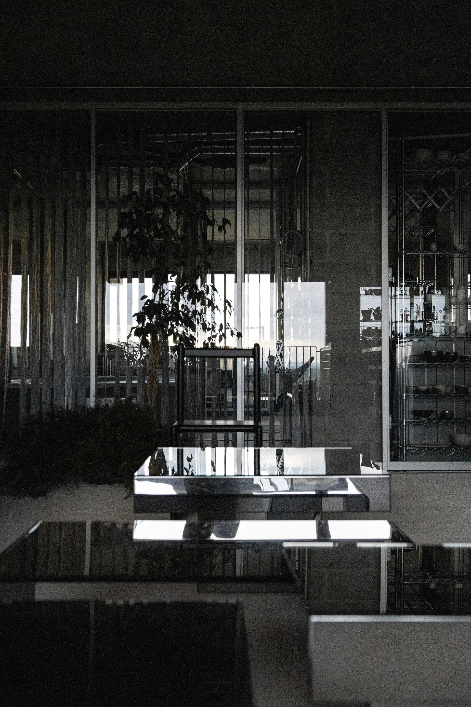A dark minimalist interior featuring highly reflective glossy tabletops in the foreground, a black-framed chair, and glass wall partitions.