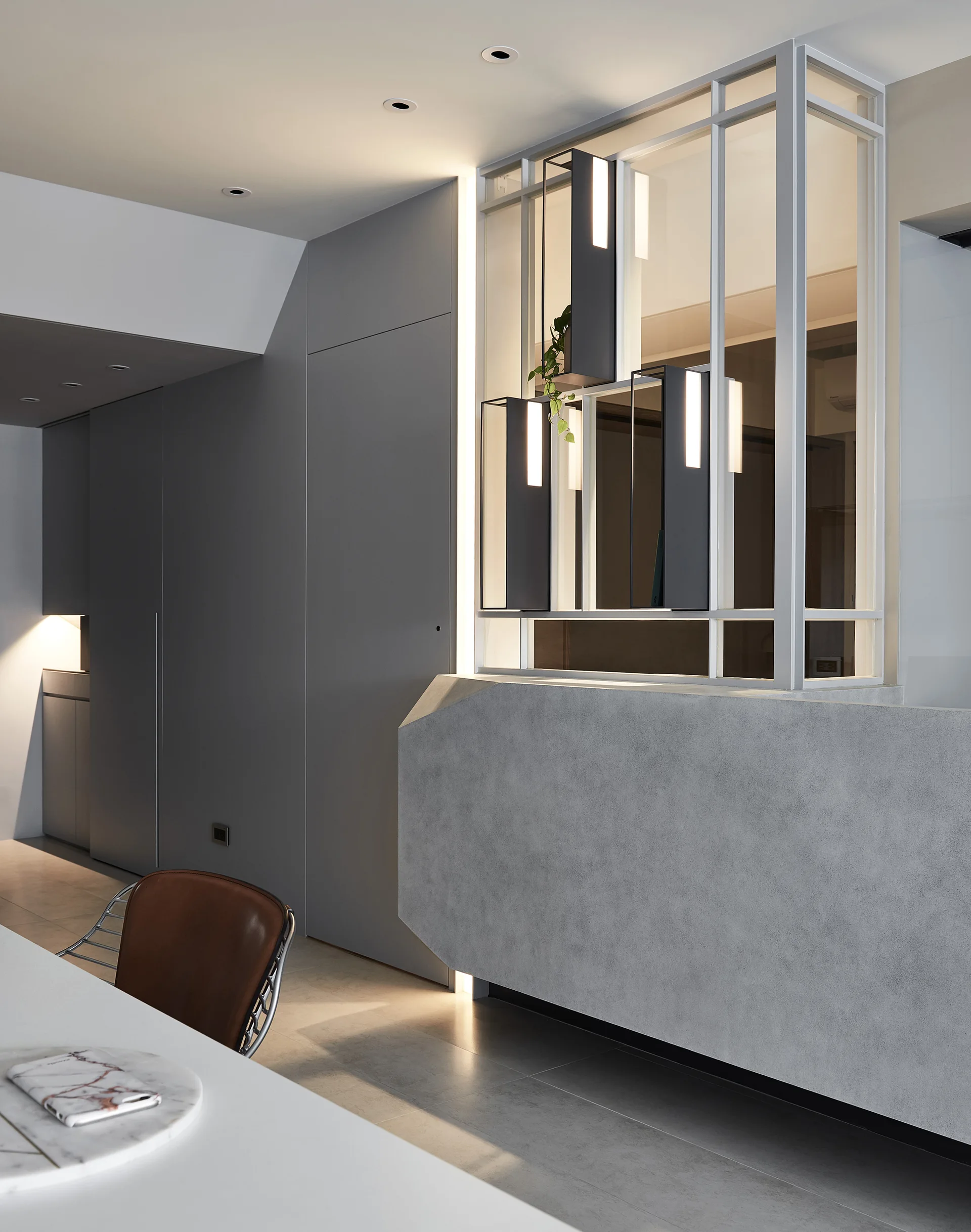 A minimalist room divider with a textured concrete base and an open metal frame containing illuminated vertical fixtures next to a brown chair.