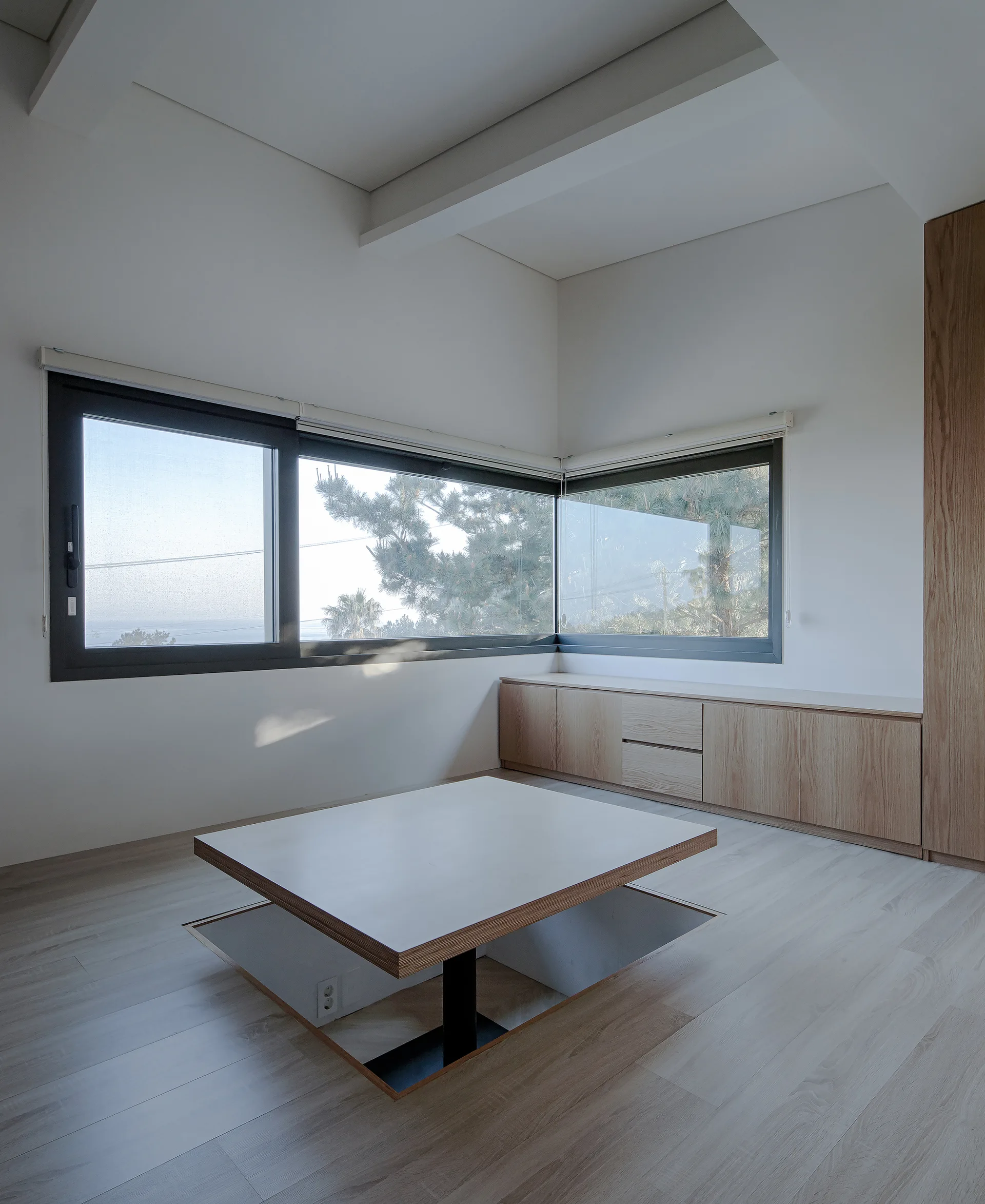 A minimalist room with a recessed floor table, low wood built-in cabinets, and large black-framed corner windows.