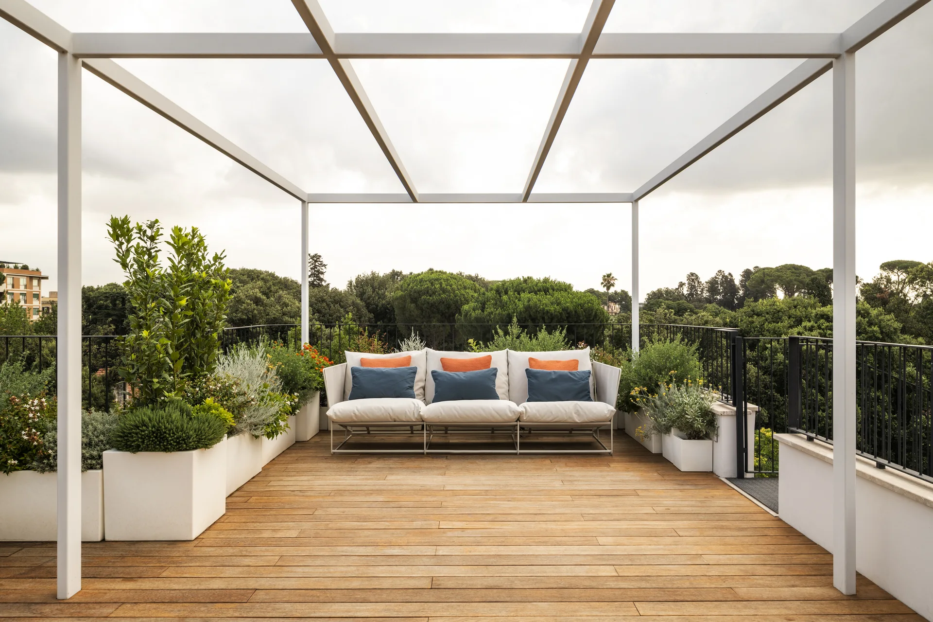 A rooftop terrace features a white metal pergola over wood decking, a white sofa with blue and orange pillows, and rectangular planters.
