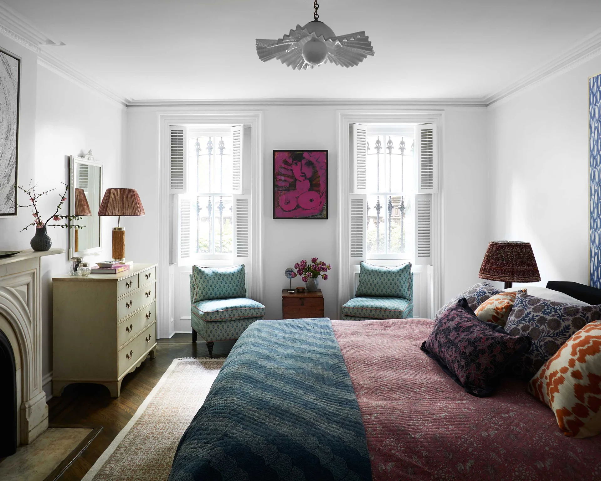 An eclectic bedroom featuring a bed with patterned bedding, a cream dresser, and two patterned armchairs placed symmetrically under shuttered windows.