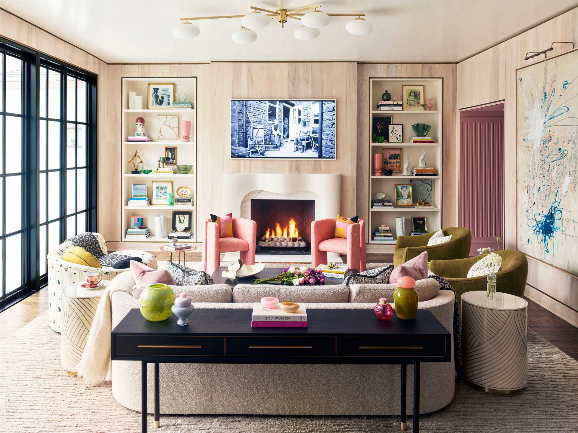 A living room featuring light wood-paneled walls, symmetrical built-in shelving flanking a marble fireplace, a curved beige sofa, and pink armchairs.