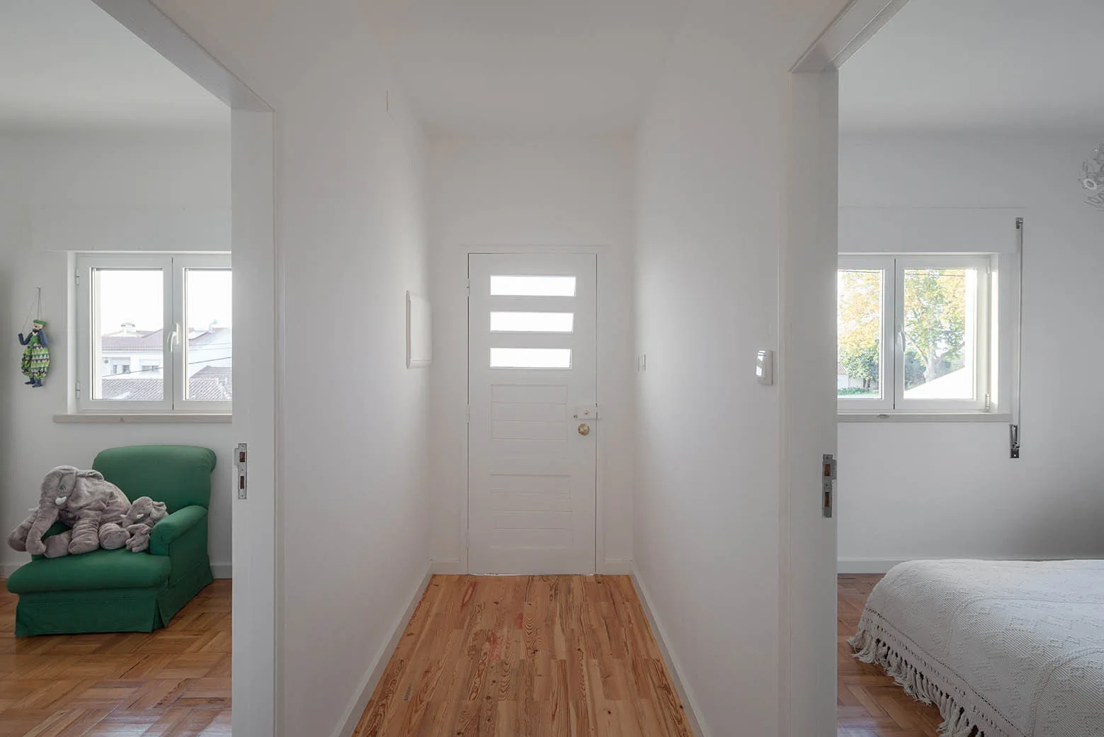 A minimalist white hallway with natural wood flooring looking toward a central entry door with open doorways on either side.