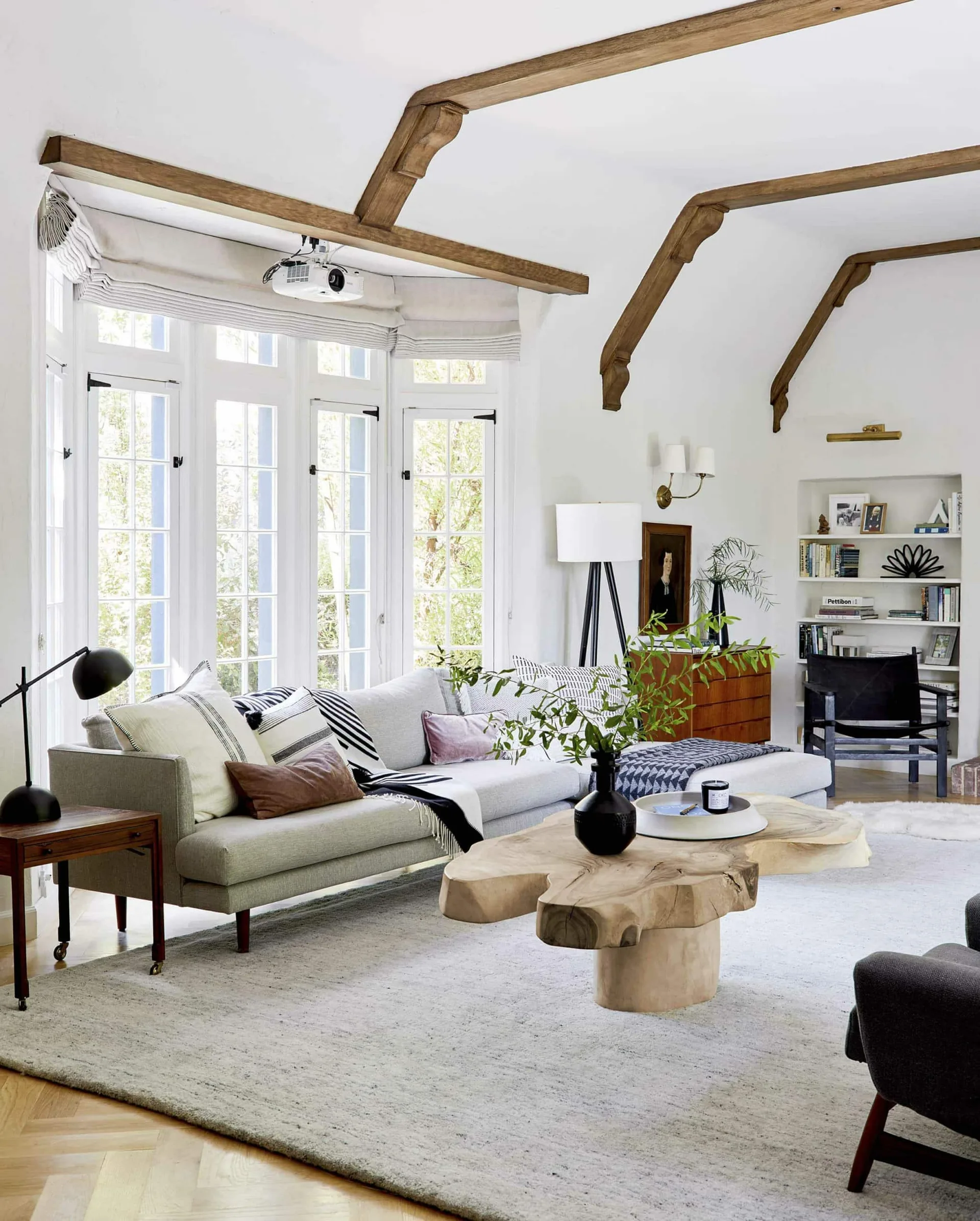 A light grey sectional sofa rests on a large area rug under a vaulted ceiling with exposed wooden beams, adjacent to a live-edge coffee table.