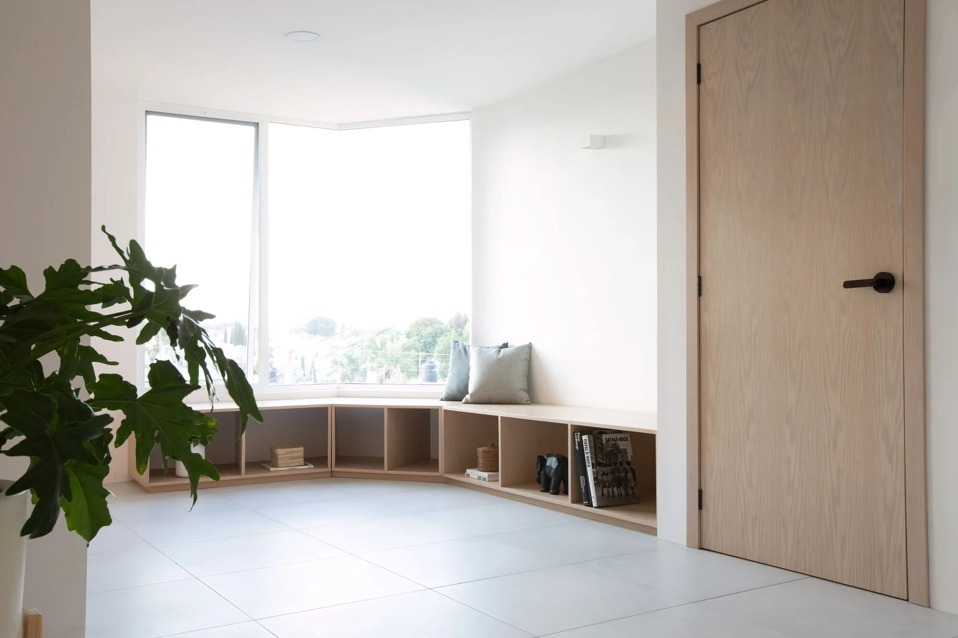 A bright minimalist room features a low light-wood cubby storage unit wrapping under a large corner window beside a matching wood door.