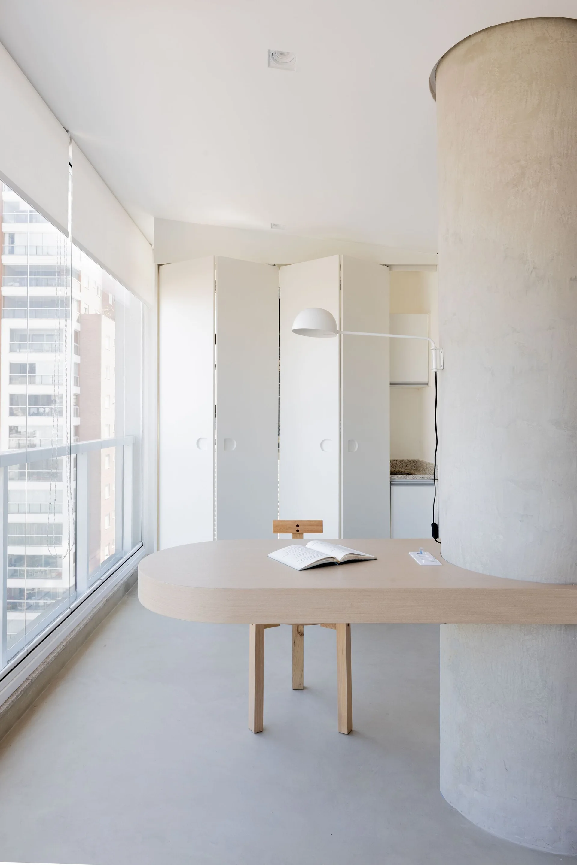 A curved light wood table is anchored to a grey concrete column in a minimalist apartment next to floor-to-ceiling windows.