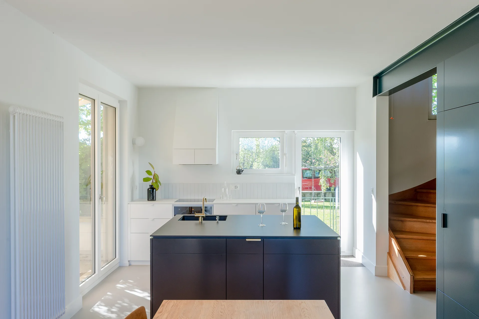 A minimalist kitchen features a dark gray island with a gold faucet, white cabinetry, and a wooden staircase visible in the background.