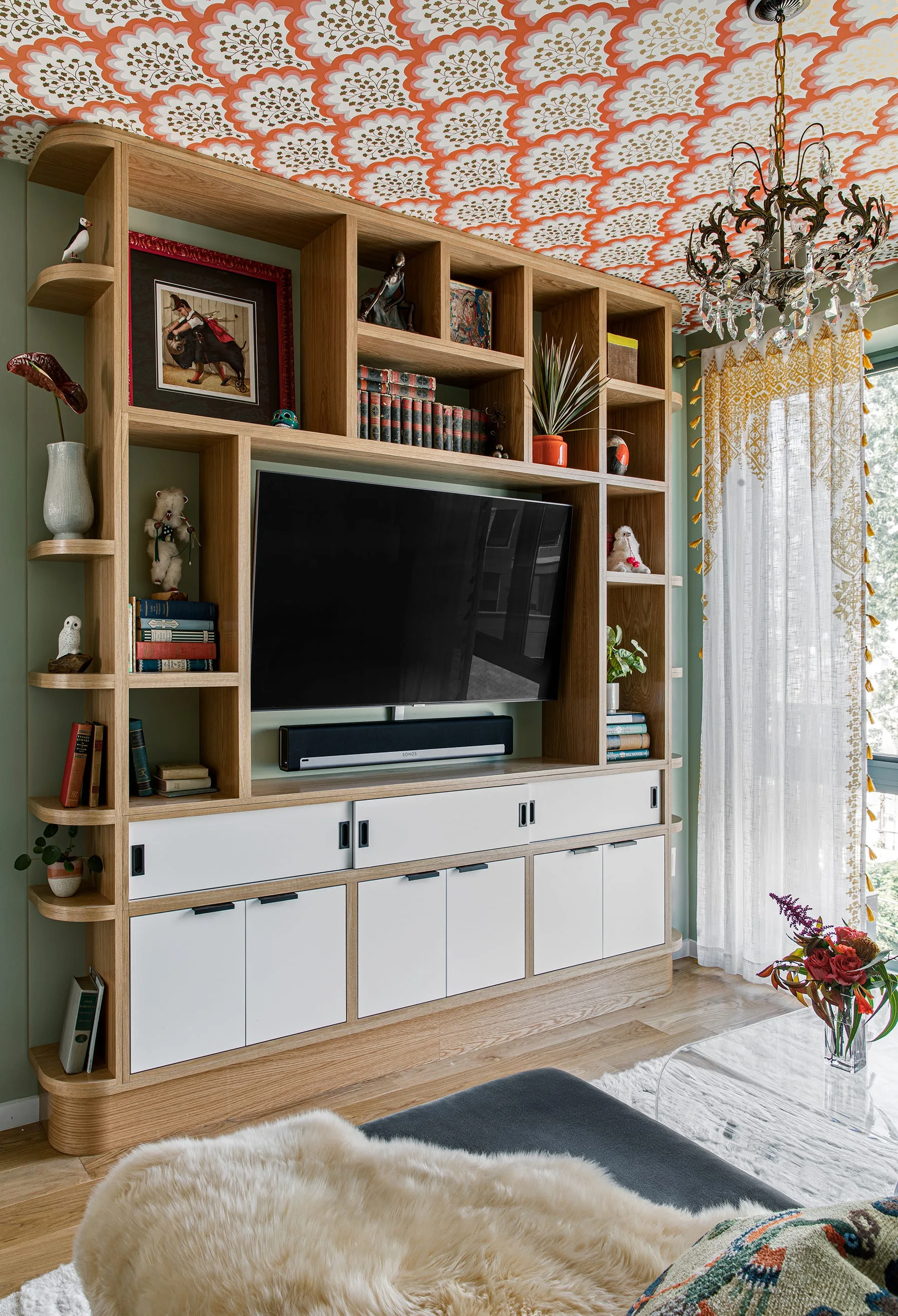 A custom light wood media unit with white cabinets is built against a sage green wall, below a ceiling covered in orange and white floral wallpaper.