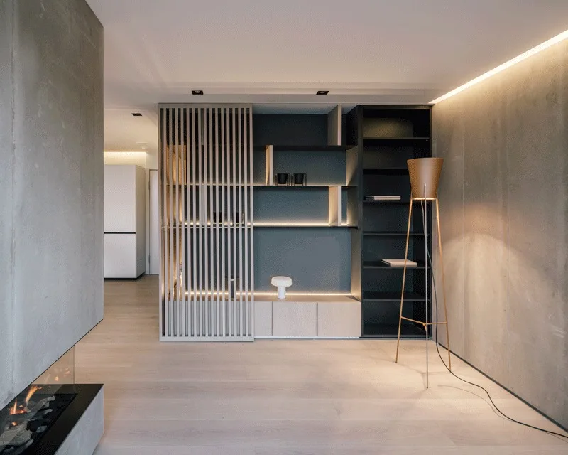 A minimalist living room features concrete walls, light wood floors, a dark built-in shelving unit with warm LED backlighting, and a wooden slatted screen.