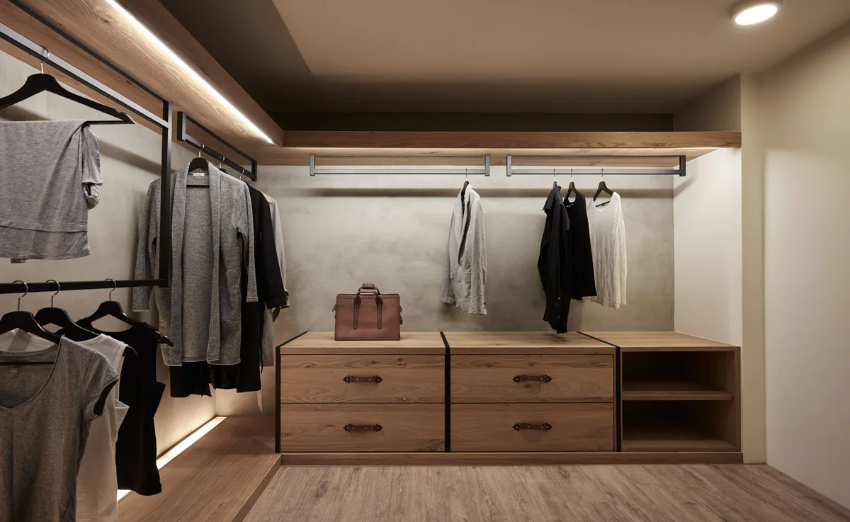 A modern walk-in closet featuring textured gray concrete walls, natural wood drawers with leather pulls, and black metal hanging garment racks.