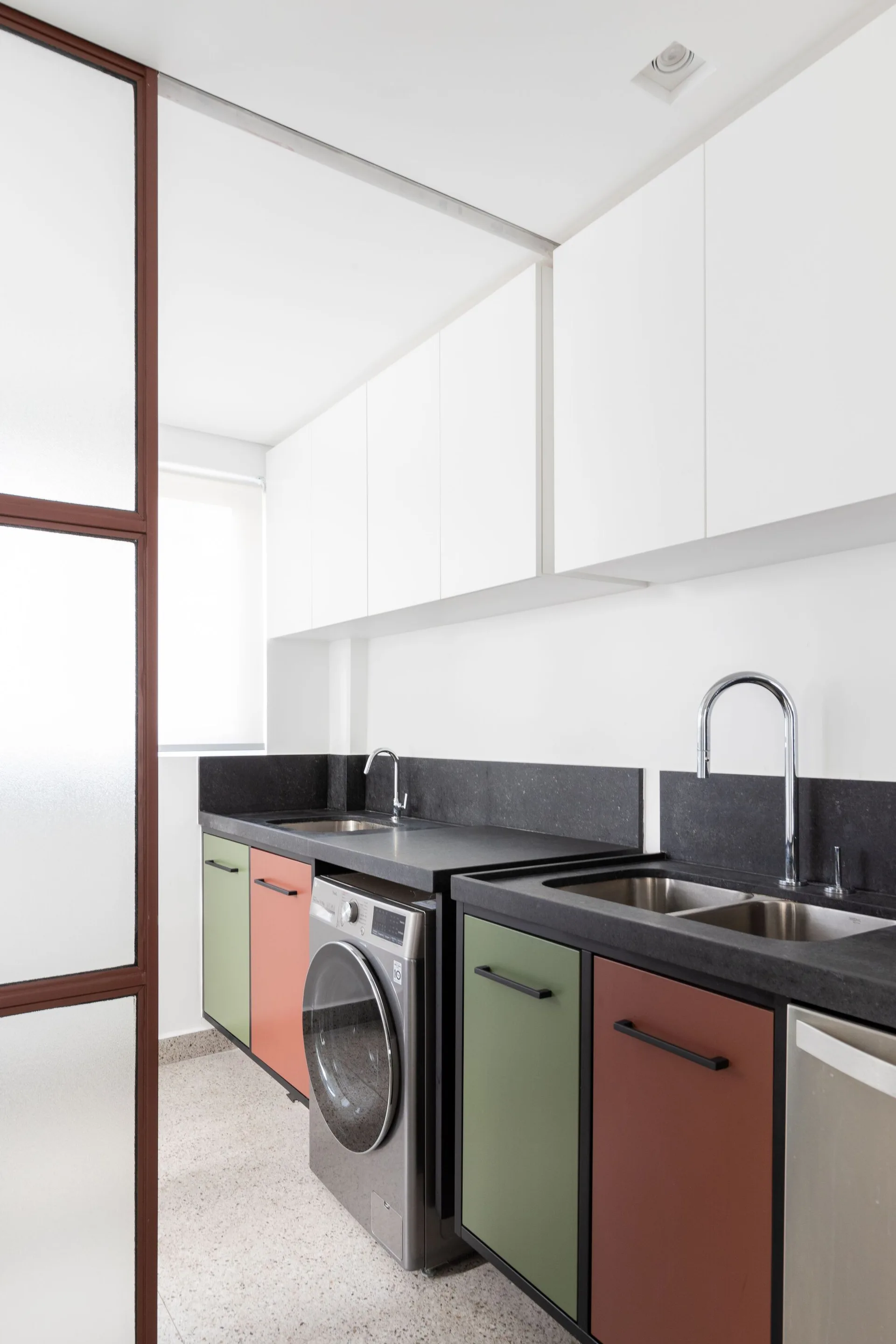 A modern utility room featuring sage green and terracotta cabinets under dark countertops next to a front-loading washer.