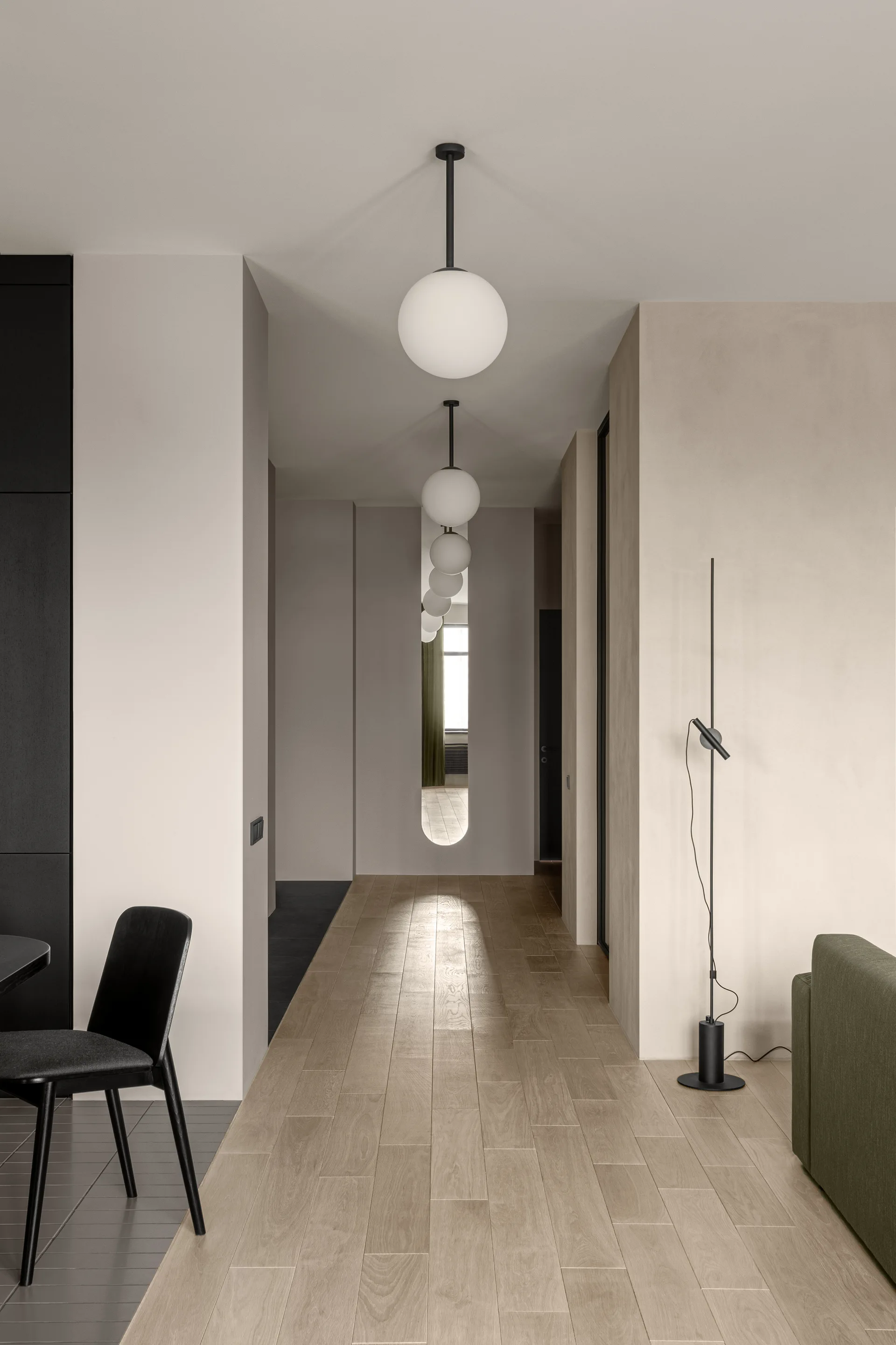 A minimalist hallway features light wood plank flooring, beige walls, a series of white globe pendant lights, and a slender black floor lamp.