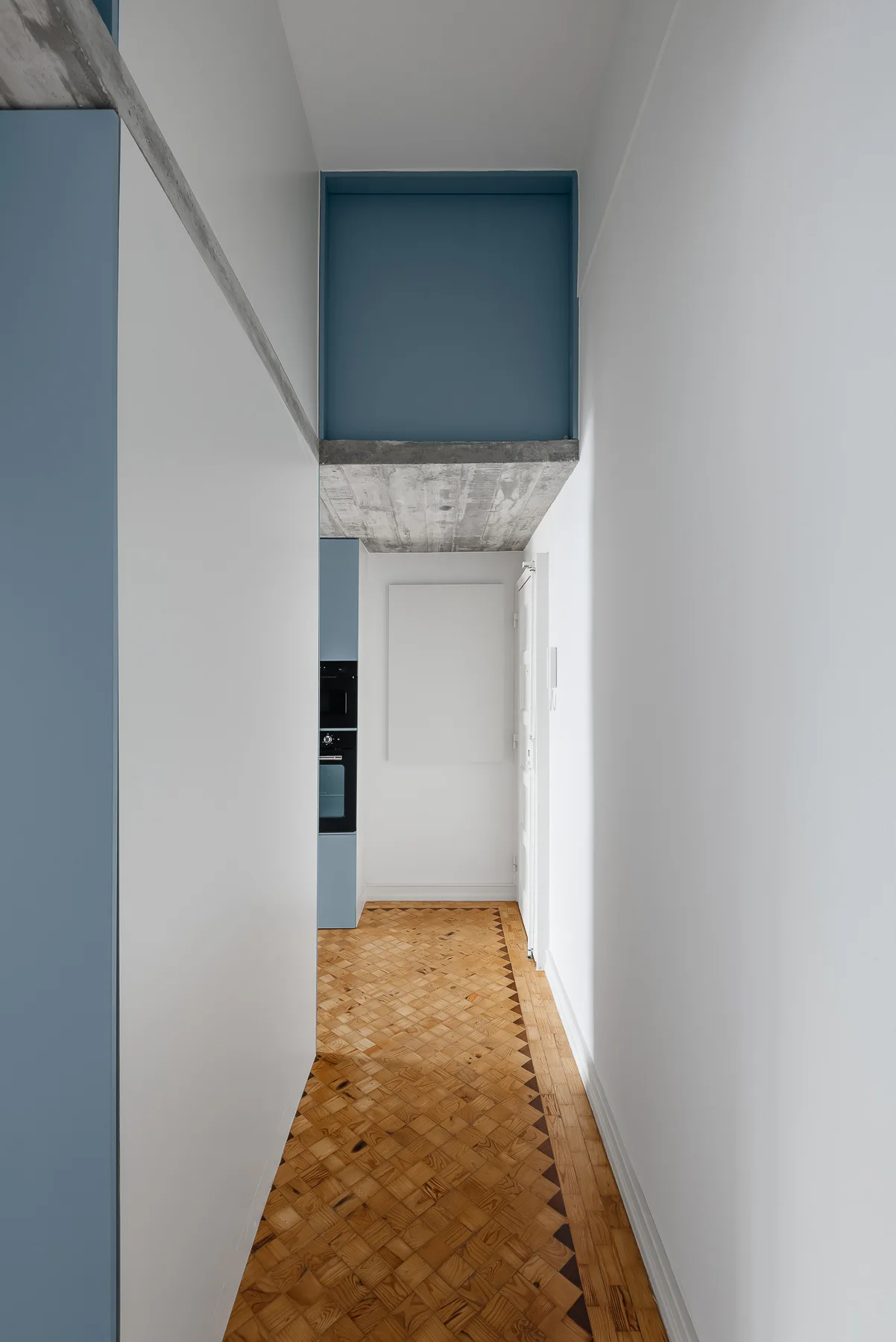 A narrow white hallway features intricate wood parquet flooring, matte blue built-in cabinetry, and an exposed concrete ceiling beam.