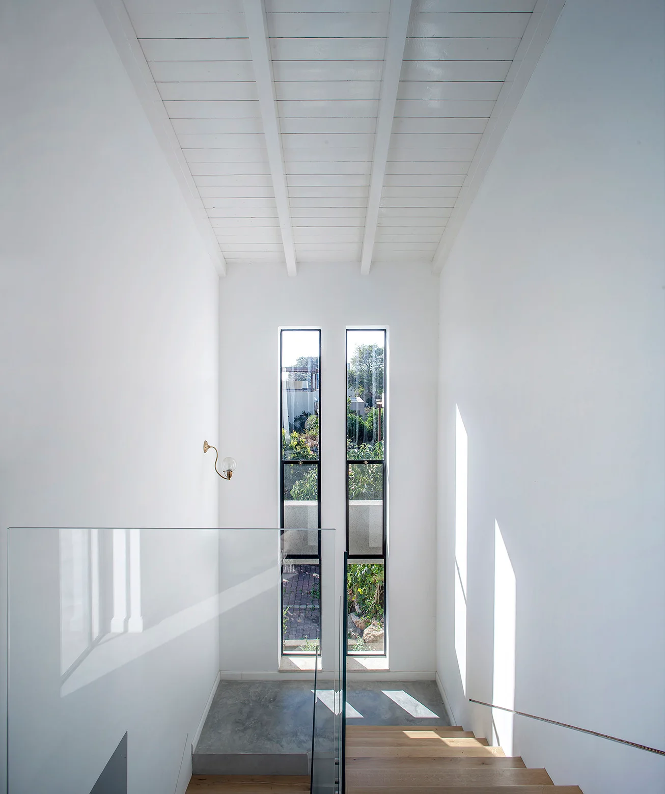 A minimalist stairwell features wooden steps, a glass railing, matte white walls, and two narrow vertical windows.