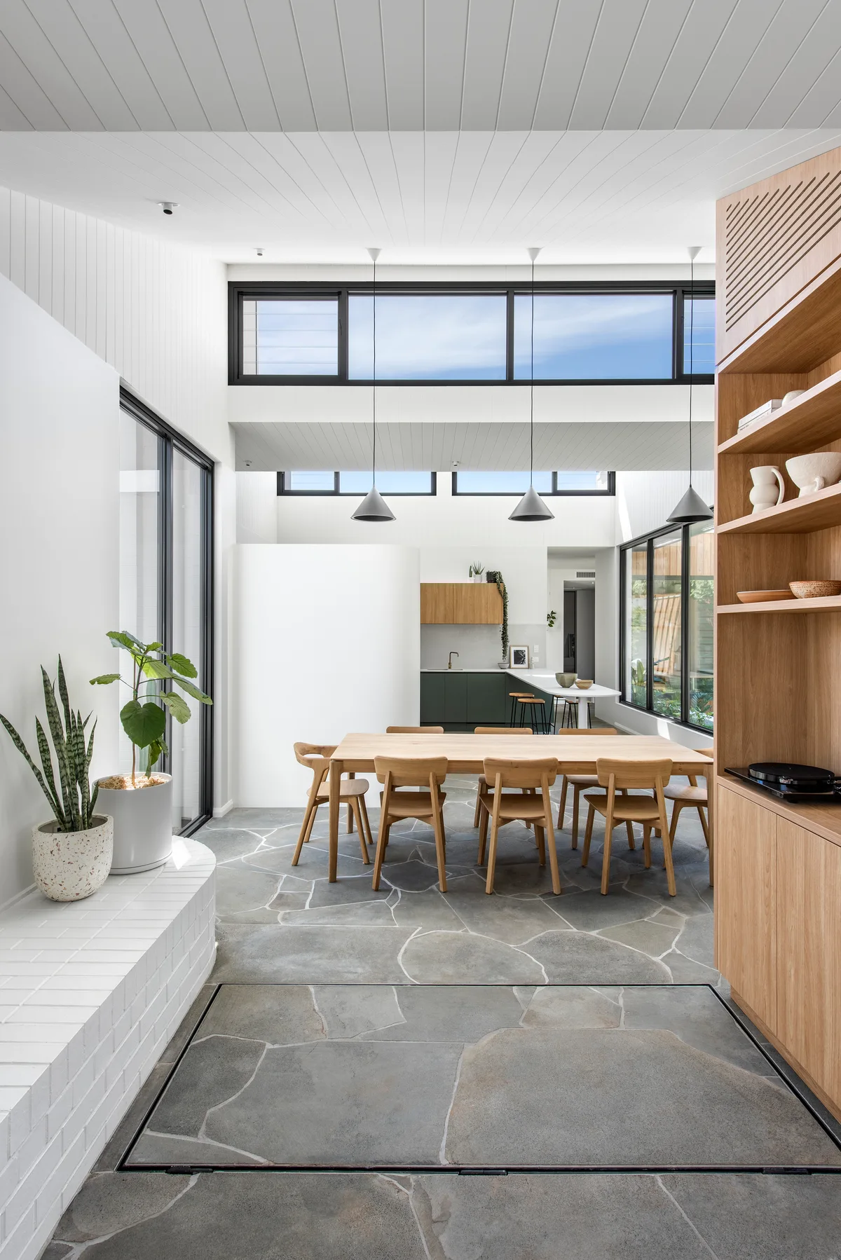 A modern dining room features a light wood table and chairs on a grey flagstone floor beneath a high ceiling with clerestory windows.