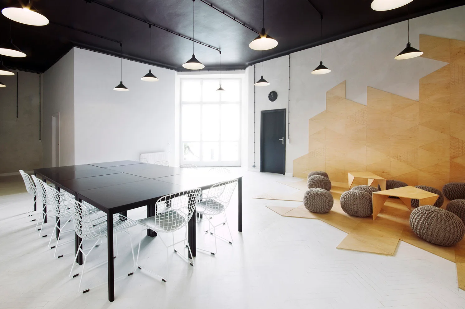 A minimalist workspace features a black conference table alongside a geometric plywood wall feature with low seating.