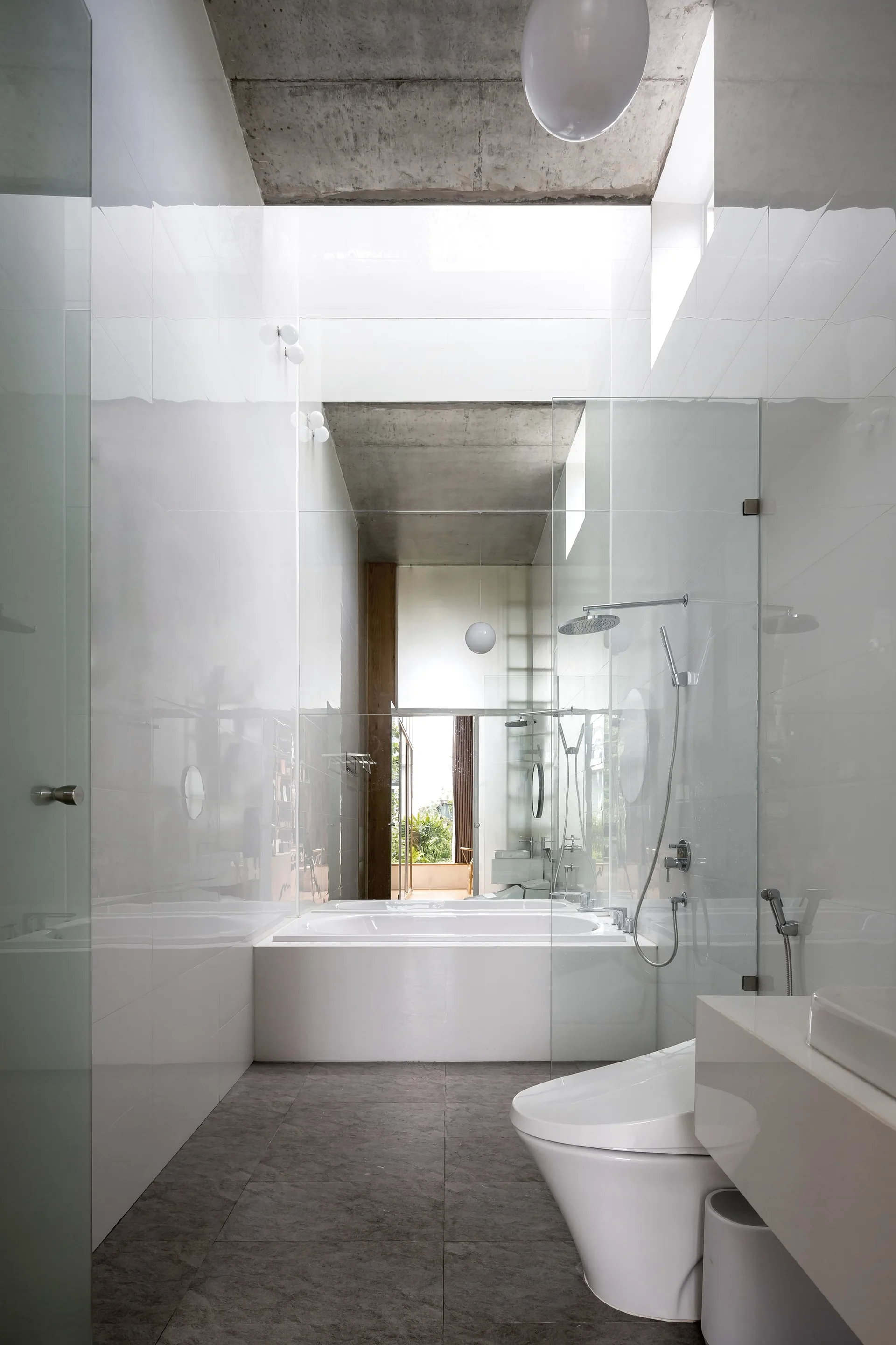 A narrow modern bathroom with glossy white tiled walls, an exposed concrete ceiling, a built-in tub, and a grey tile floor.