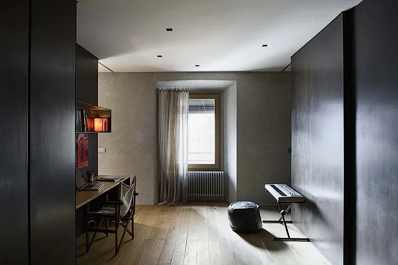 A minimalist home office features dark floor-to-ceiling wall panels, a wood desk with a chair, and a sheer curtain over a central window.