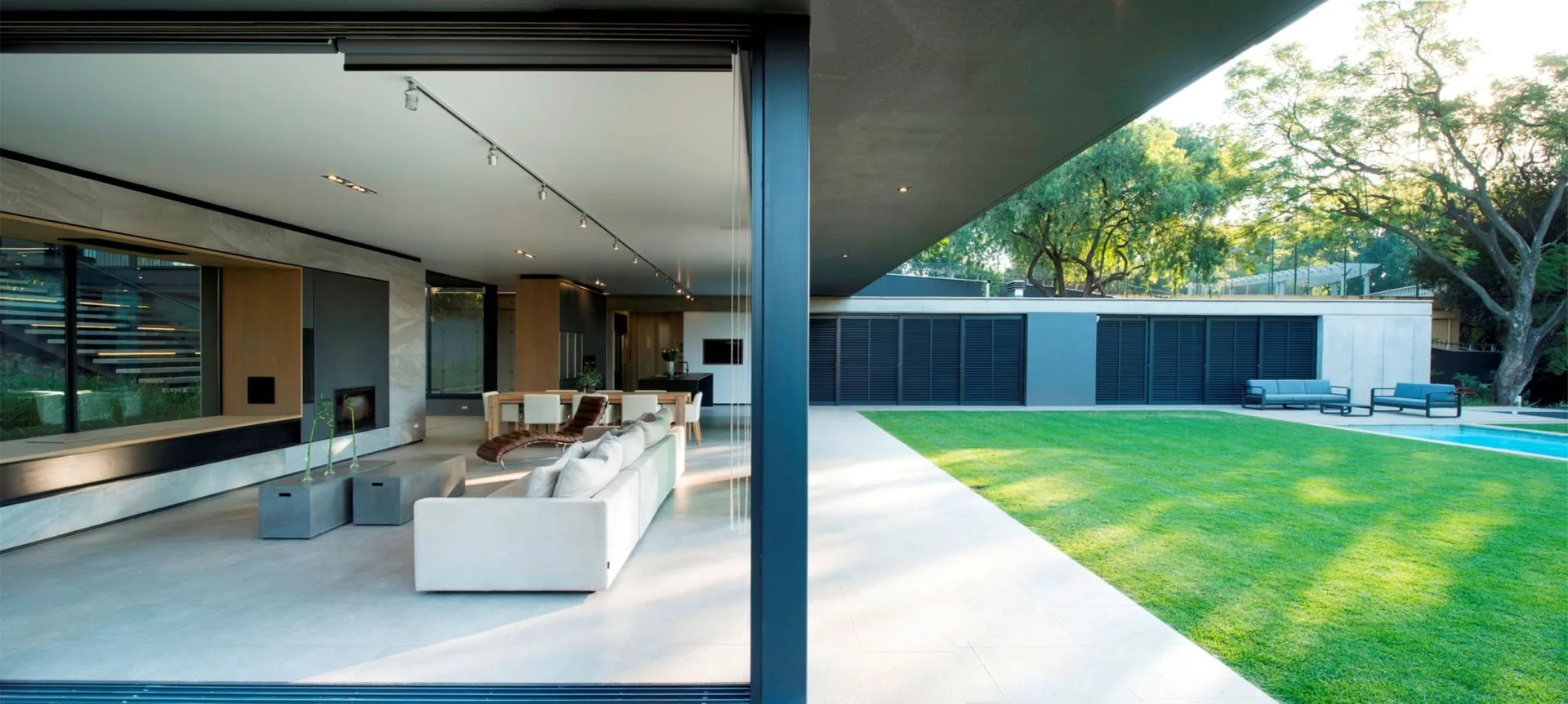 An open-concept living room with a grey sofa and concrete coffee tables features sliding glass doors that open to a manicured lawn and swimming pool.