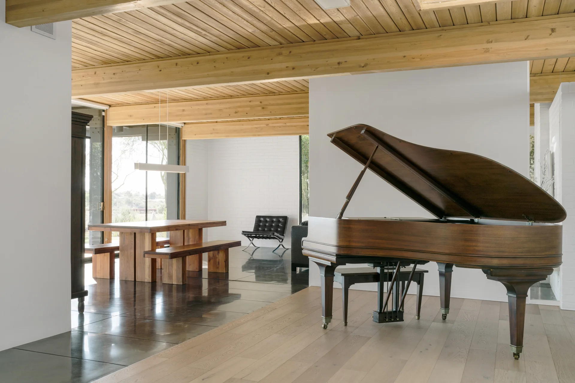 A spacious open-plan room features a brown grand piano on light wood floors and a dining table on dark polished tile under an exposed beam ceiling.
