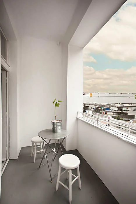 A minimalist white balcony features a grey painted floor, a small metal bistro table with a potted plant, and two white wooden stools.