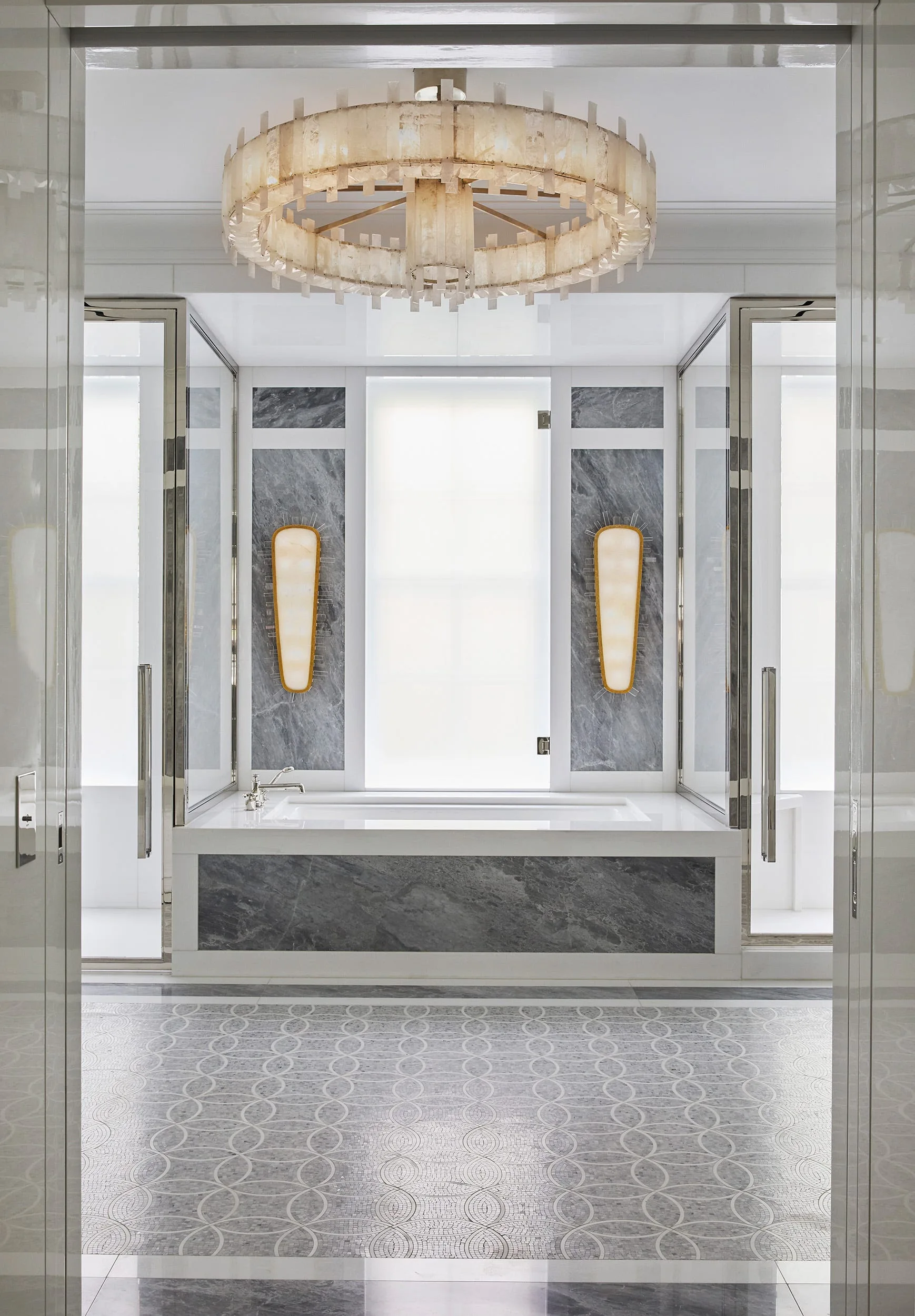 Symmetrical bathroom featuring a central marble-clad bathtub, a large circular ring chandelier, patterned stone tile flooring, and vertical wall sconces.