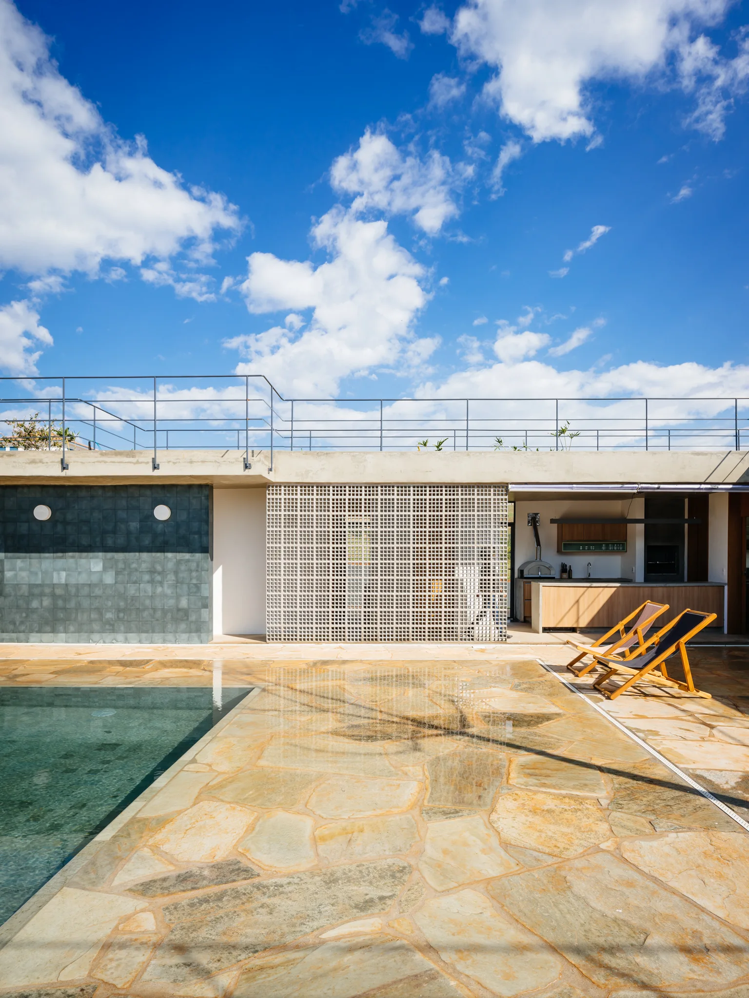 An outdoor swimming pool surrounded by a flagstone patio, a white geometric breeze block partition, and wooden lounge chairs under a blue sky.