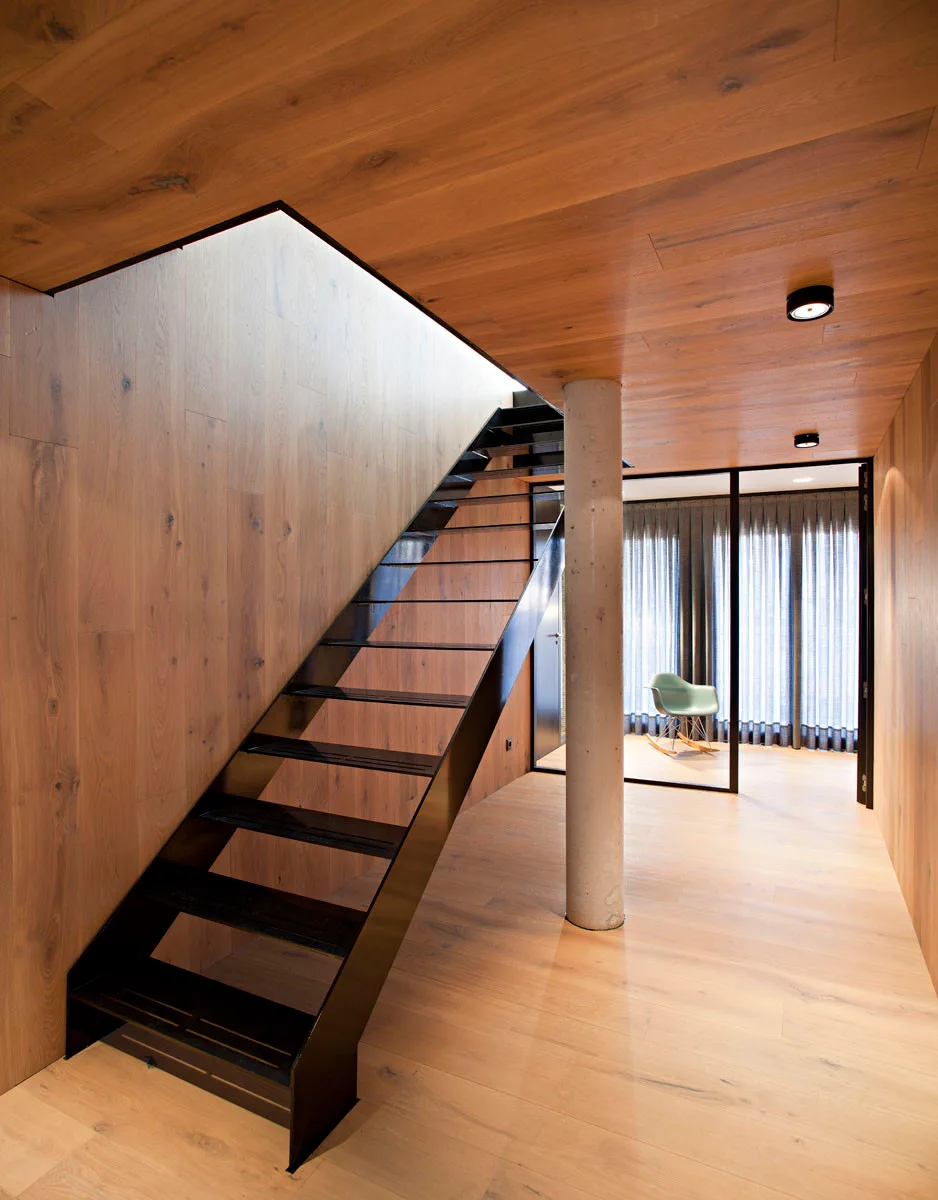 A minimalist black metal open-tread staircase rises between wood-paneled walls and ceiling, adjacent to a cylindrical concrete support column.