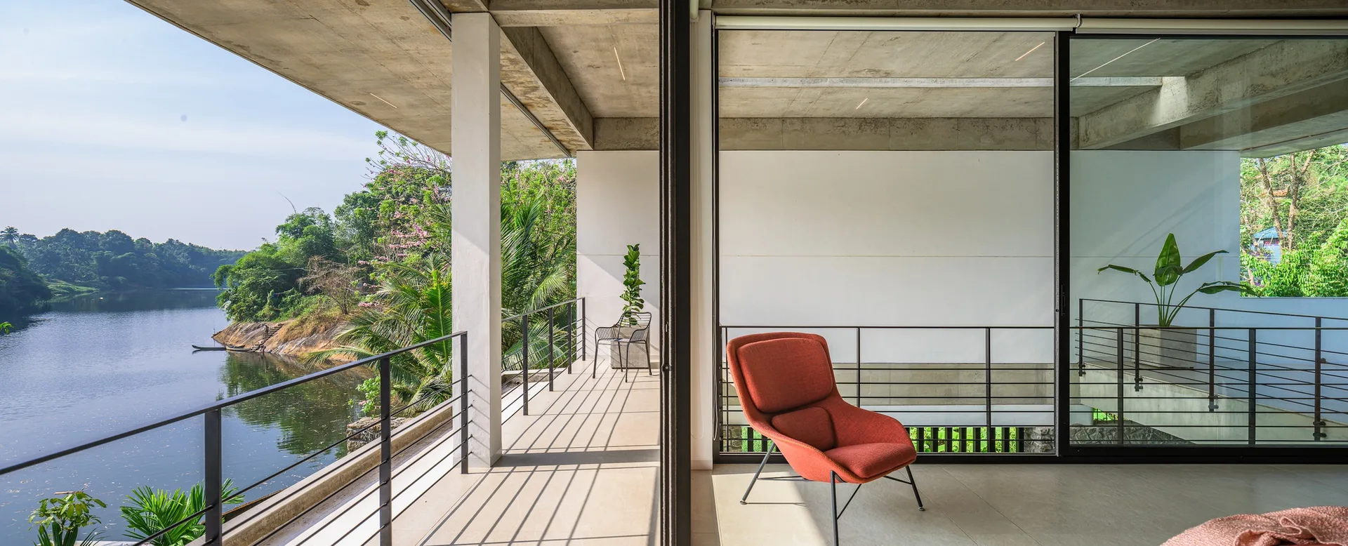A minimalist room features an orange armchair next to sliding glass doors that open to a concrete balcony overlooking a river.