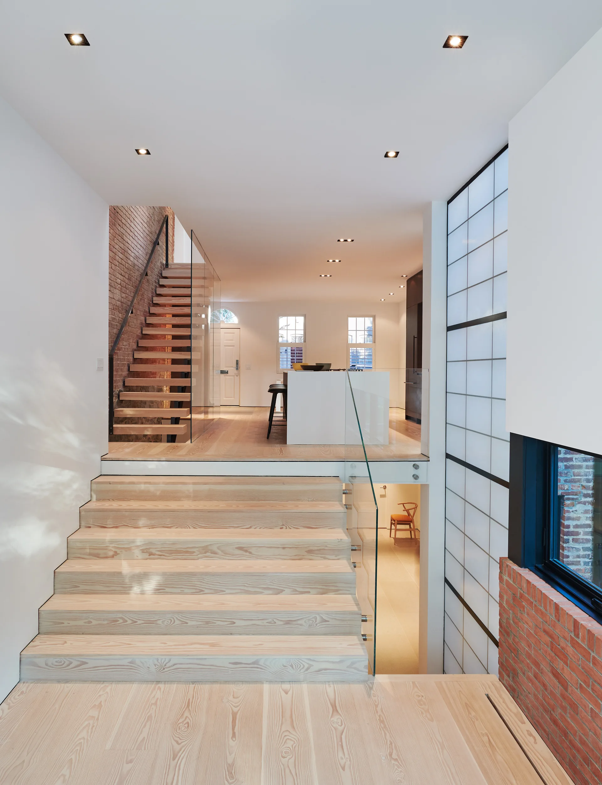 A split-level home interior featuring wide light-wood staircases, frameless glass railings, an exposed brick wall, and a view into an open kitchen.