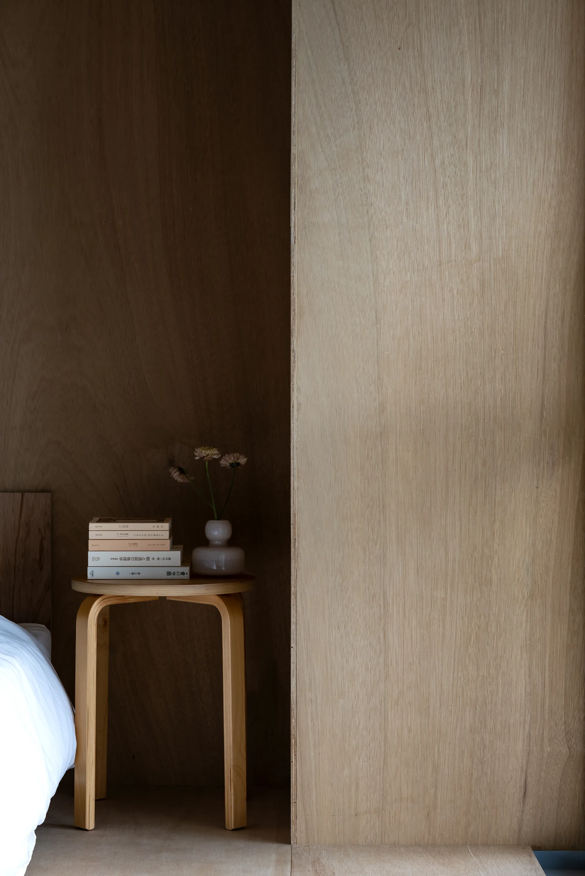 A light wood bentwood stool serves as a nightstand against a plywood wall, holding a small stack of books and a ceramic vase with flowers.